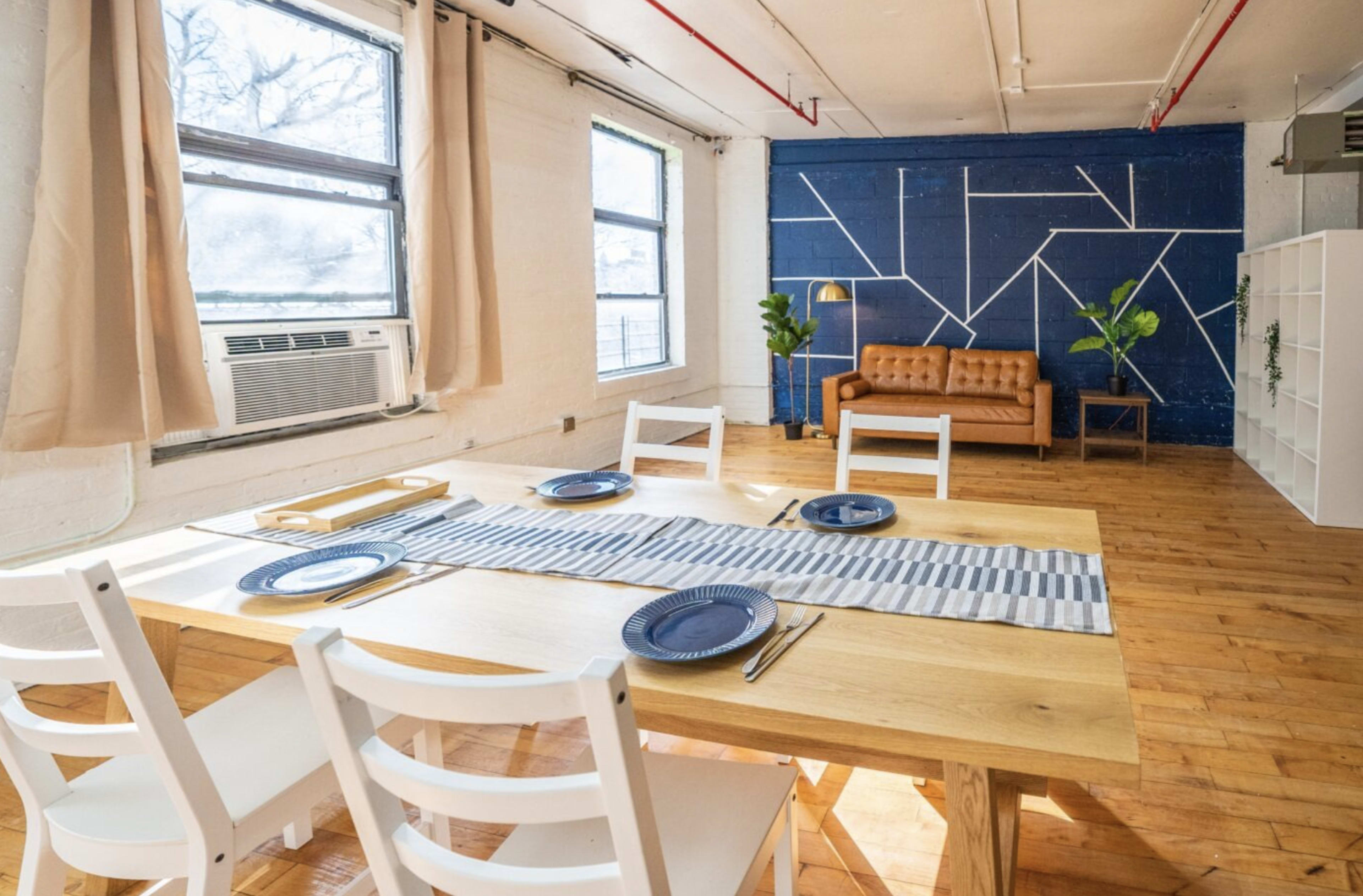 A dining area features a wooden table set for four, with chairs, a decorative table runner, and a seating area in the background that includes a brown couch and a plant against a blue accent wall.