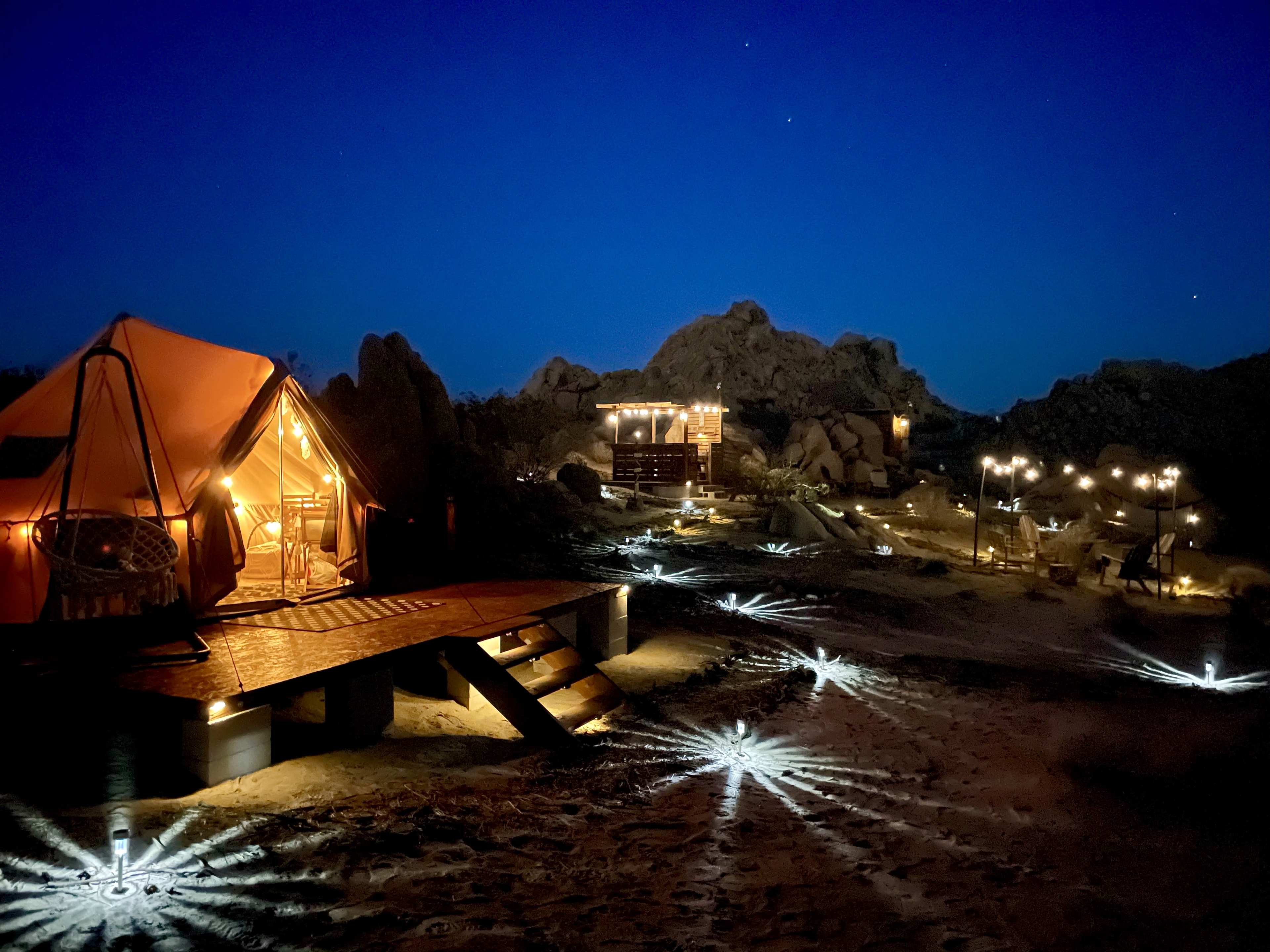 A tent illuminated by warm light stands on a wooden platform among rocky formations, with ground lights creating star-like patterns in the sand.