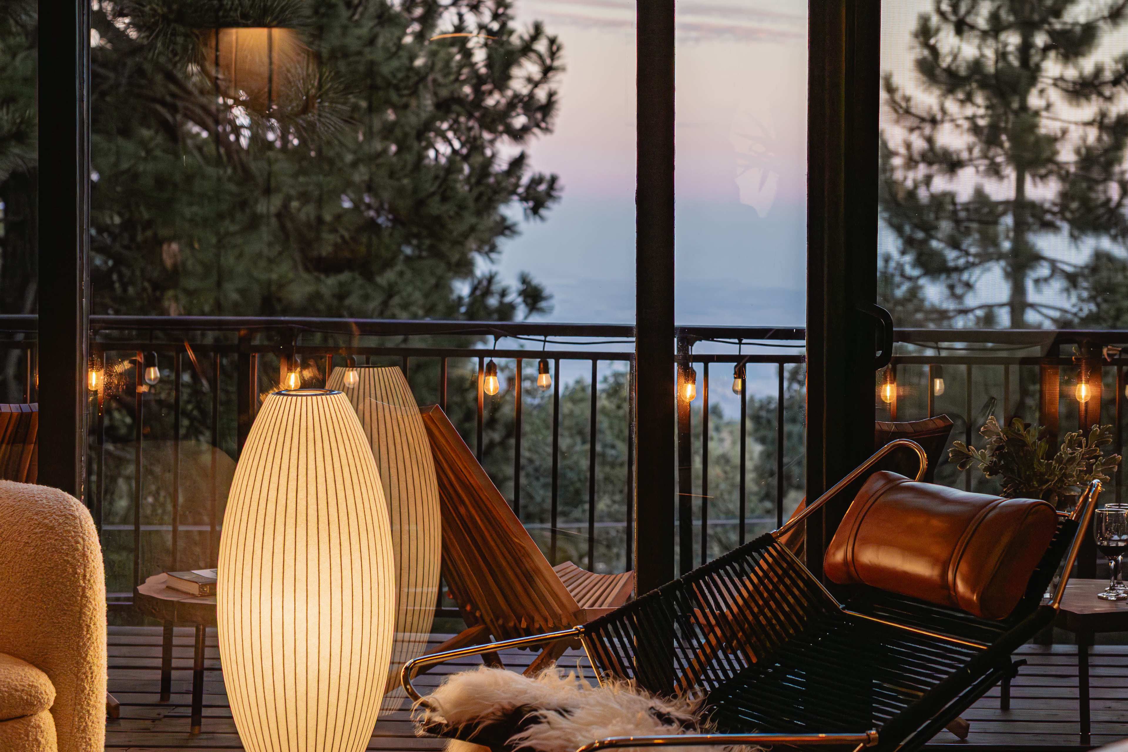 A modern interior features a sleek, curved floor lamp beside a stylish lounge chair on a deck with a view of trees and a sunset.