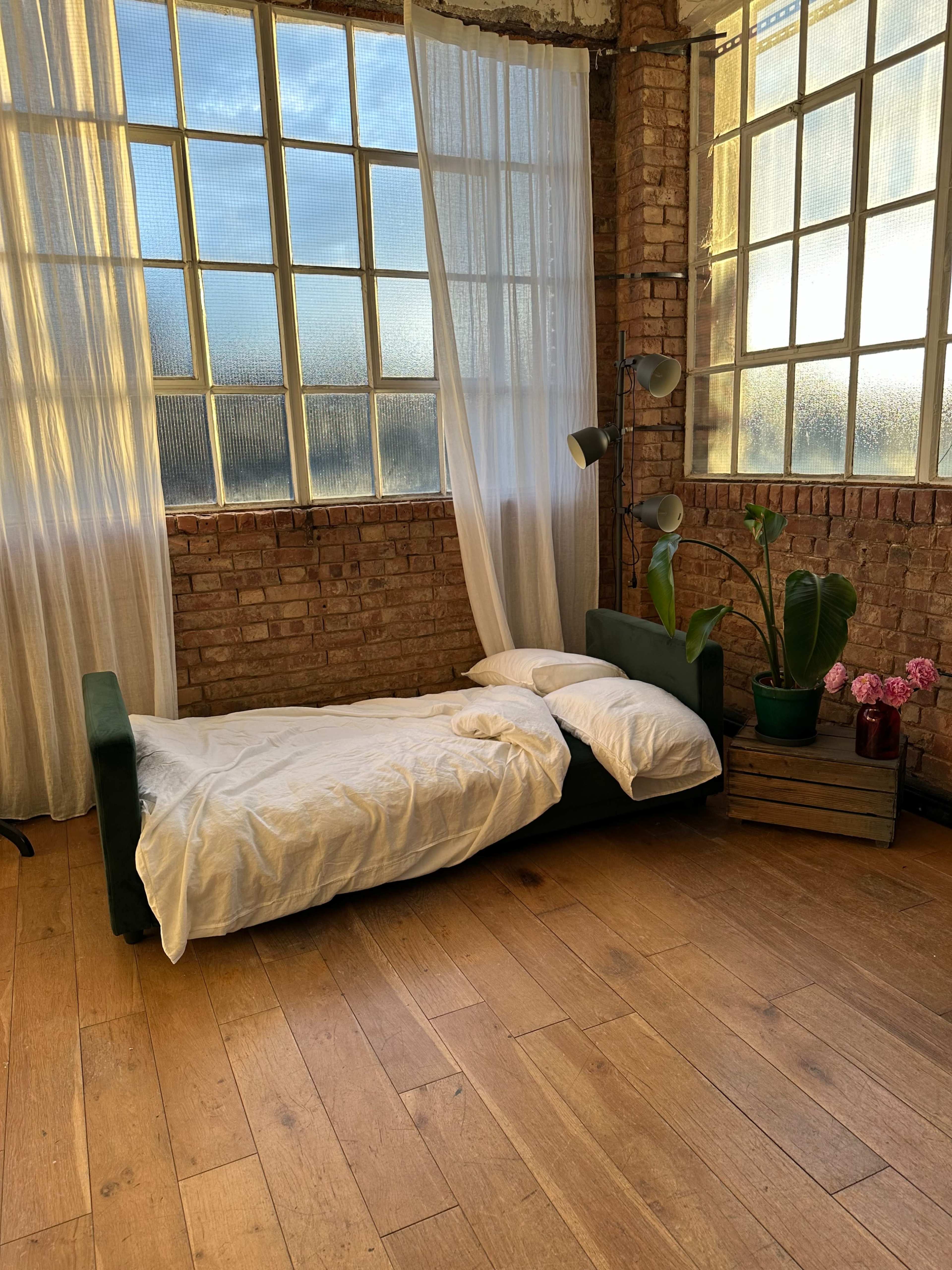 A minimalistic room with a bed made of a green frame, white bedding, large windows allowing natural light, and potted plants and flowers in the corner.