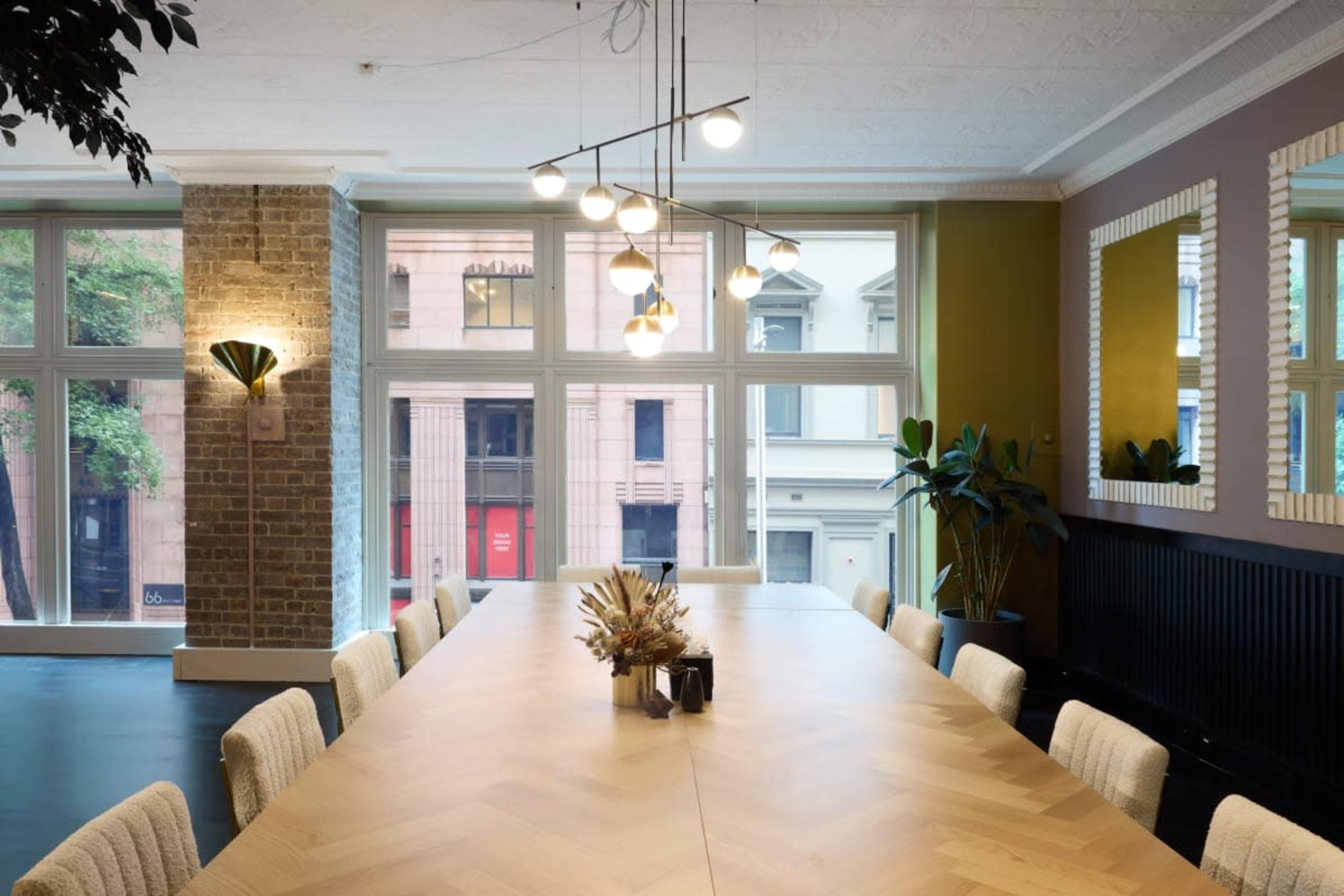 A long wooden conference table is positioned in a brightly lit room with large windows, potted plants, and modern lighting fixtures.