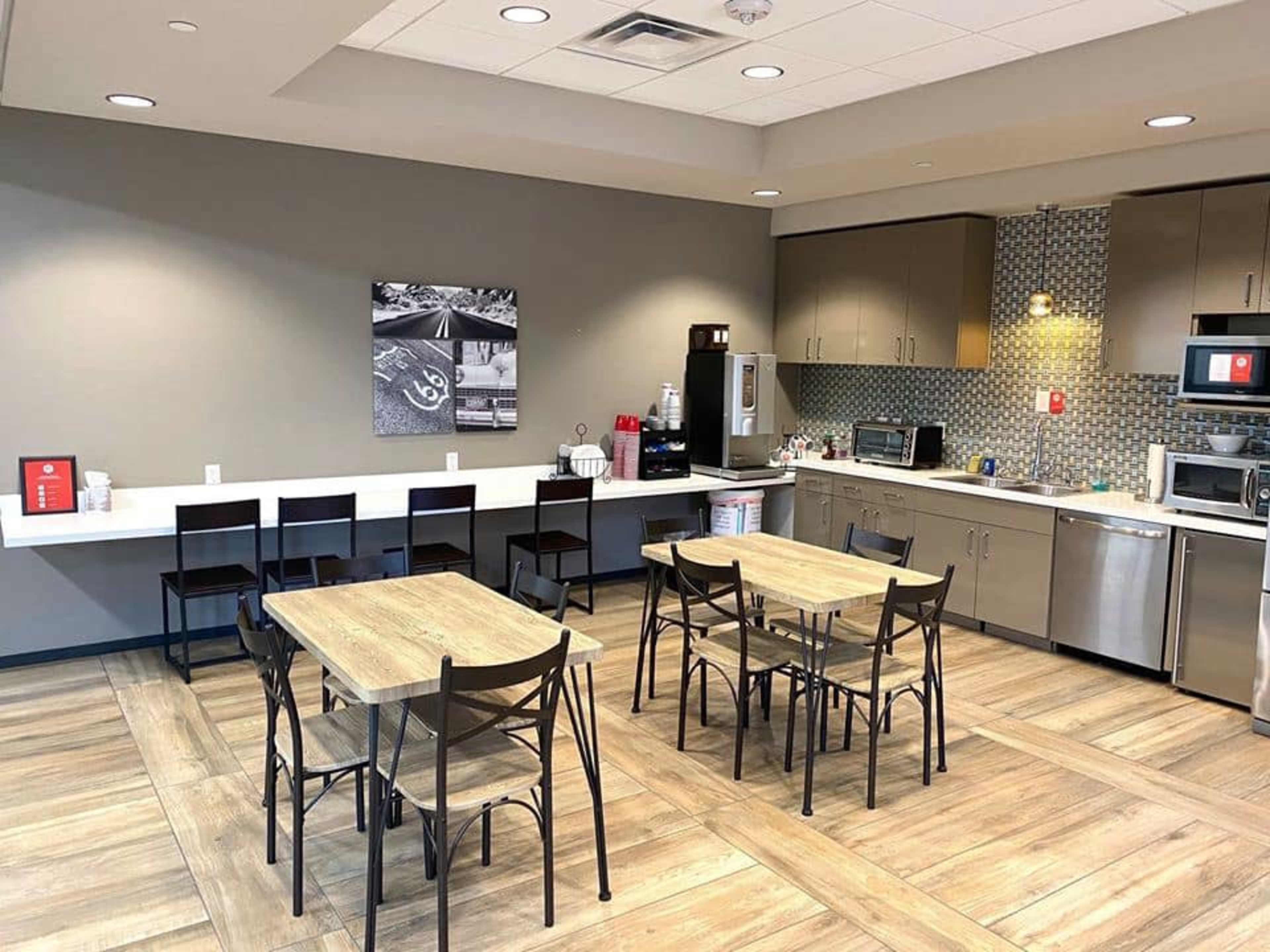 A modern kitchen area with two tables and chairs, a countertop with a coffee station, and various appliances along the wall.