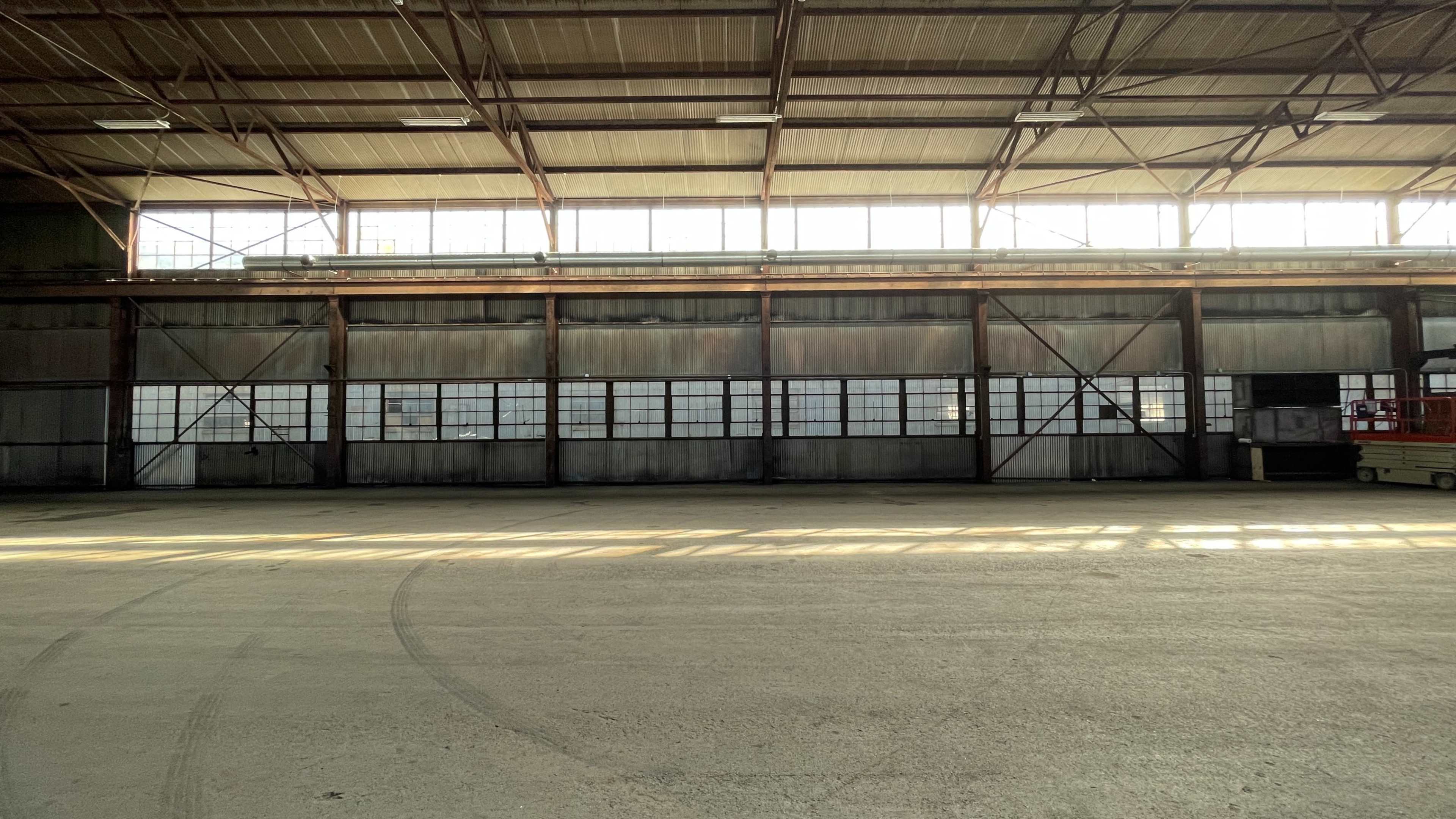 The image shows the interior of a large, empty industrial warehouse with metal beams and numerous windows allowing natural light to enter.