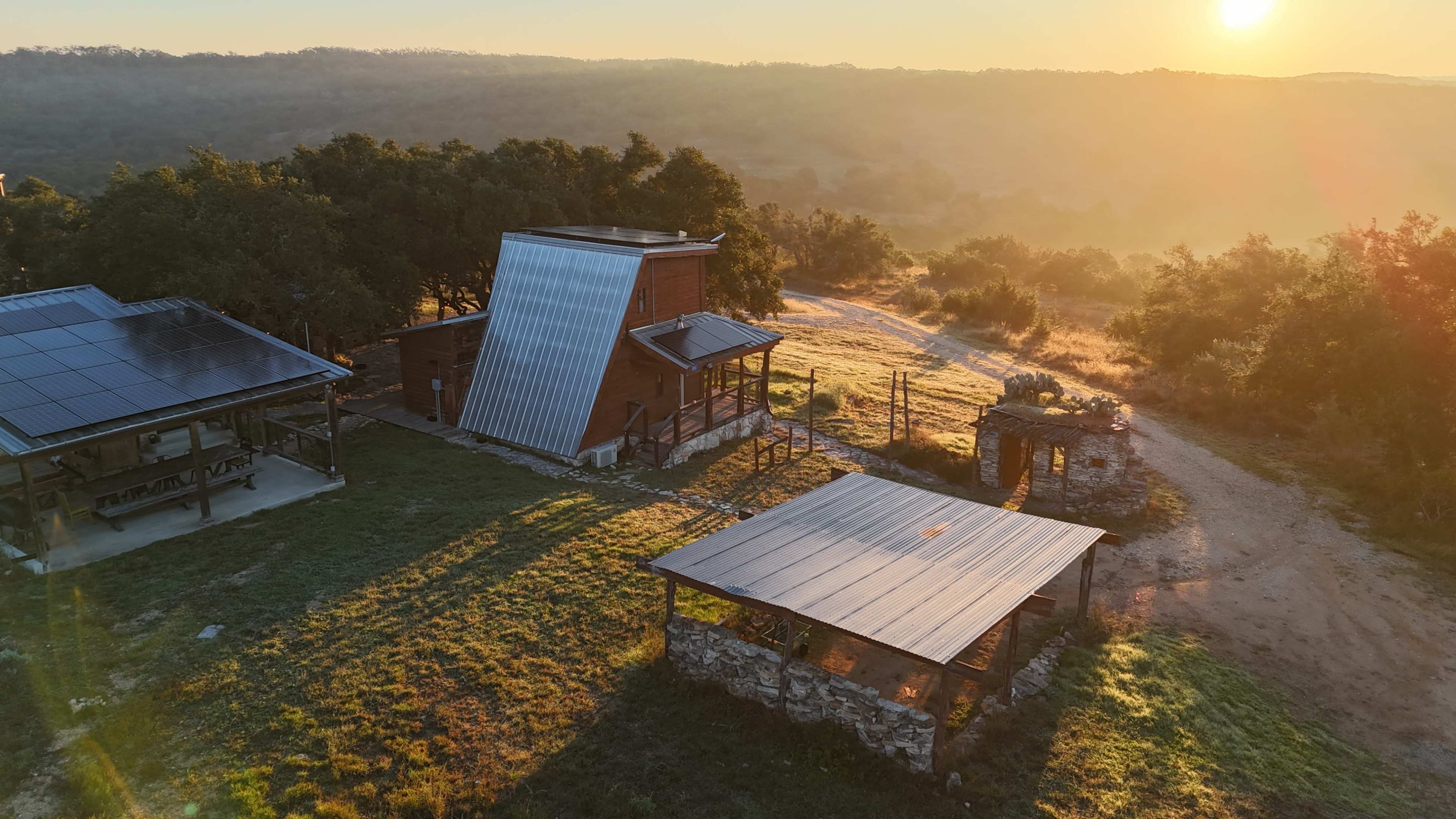 The image shows a rustic cabin with a metal roof and solar panels, surrounded by a grassy landscape bathed in warm sunlight during sunrise.