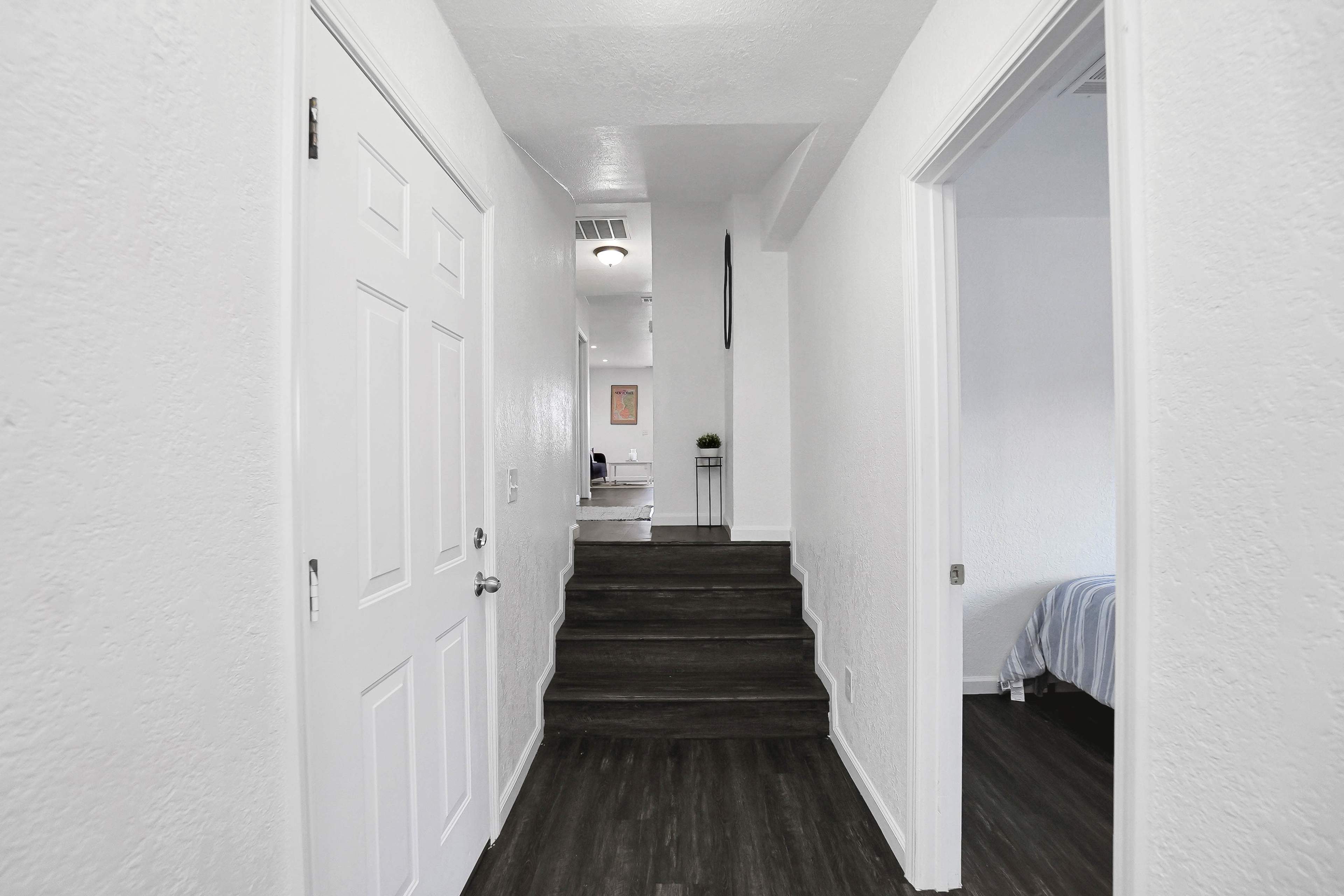A clean hallway with white walls and a dark wood floor leads towards stairs and an open space in the background.