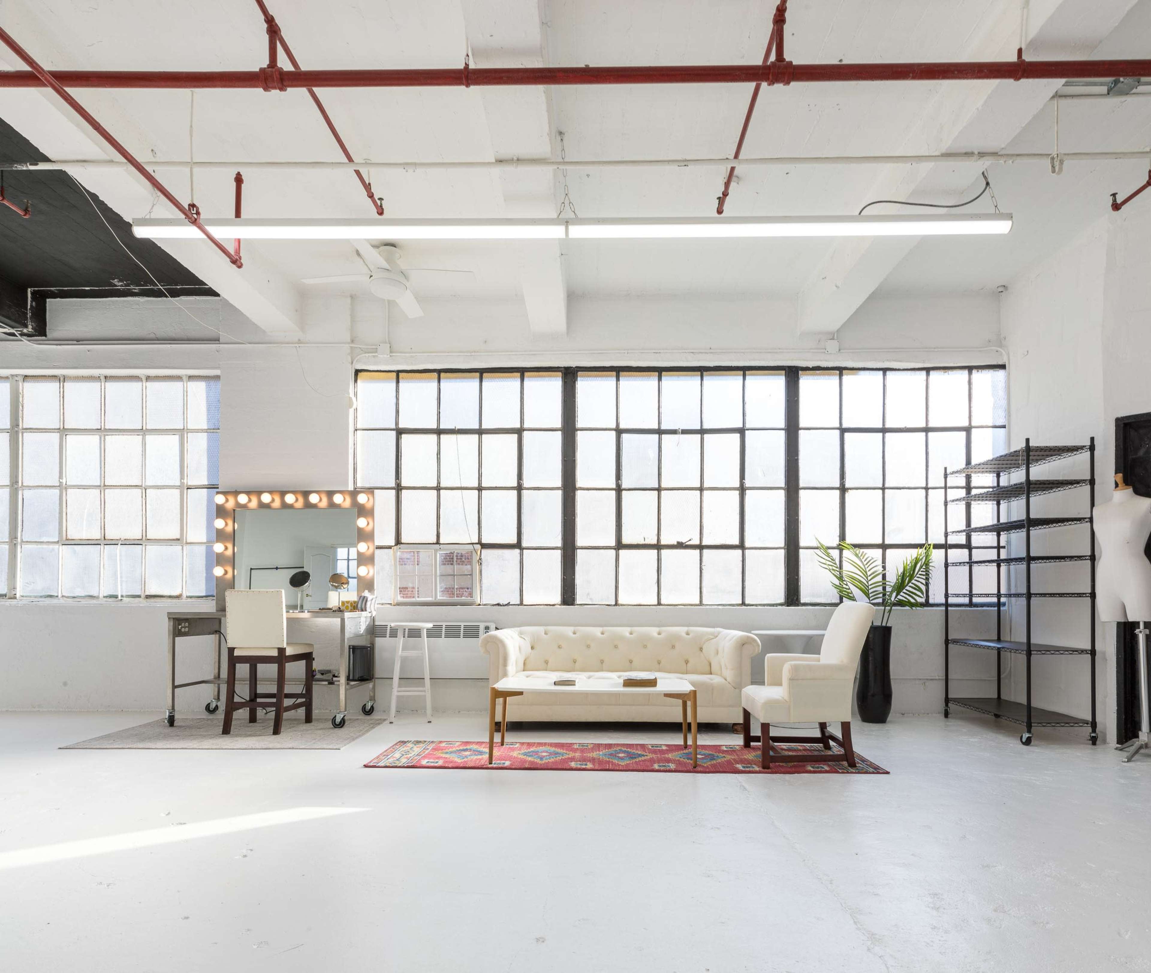 A spacious room features large windows, a tufted white sofa, a decorative mirror with lights, a metal shelving unit, and a plant.