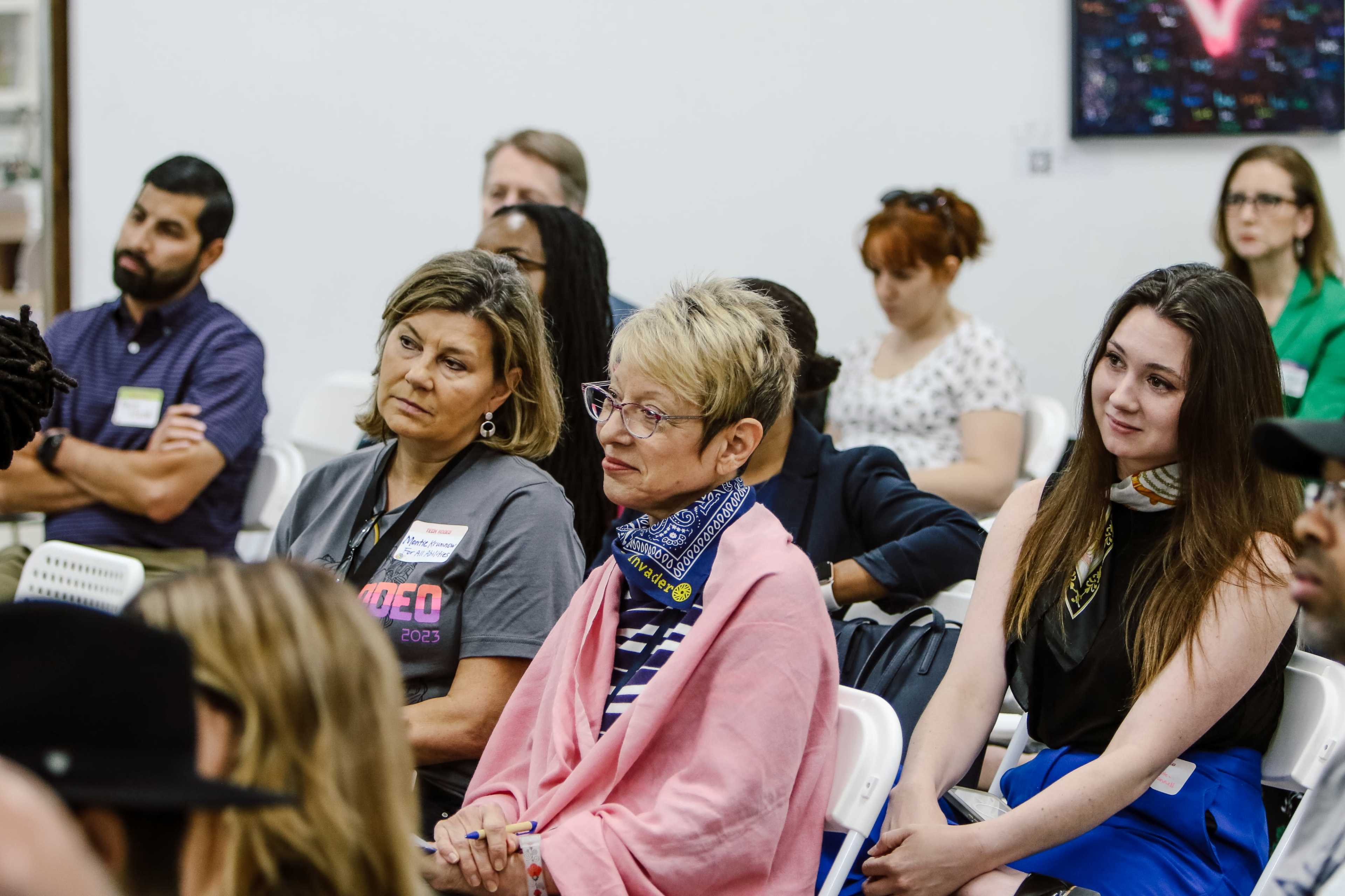 A group of people sits attentively in an indoor setting, with some wearing name tags and diverse expressions as they listen to a speaker.