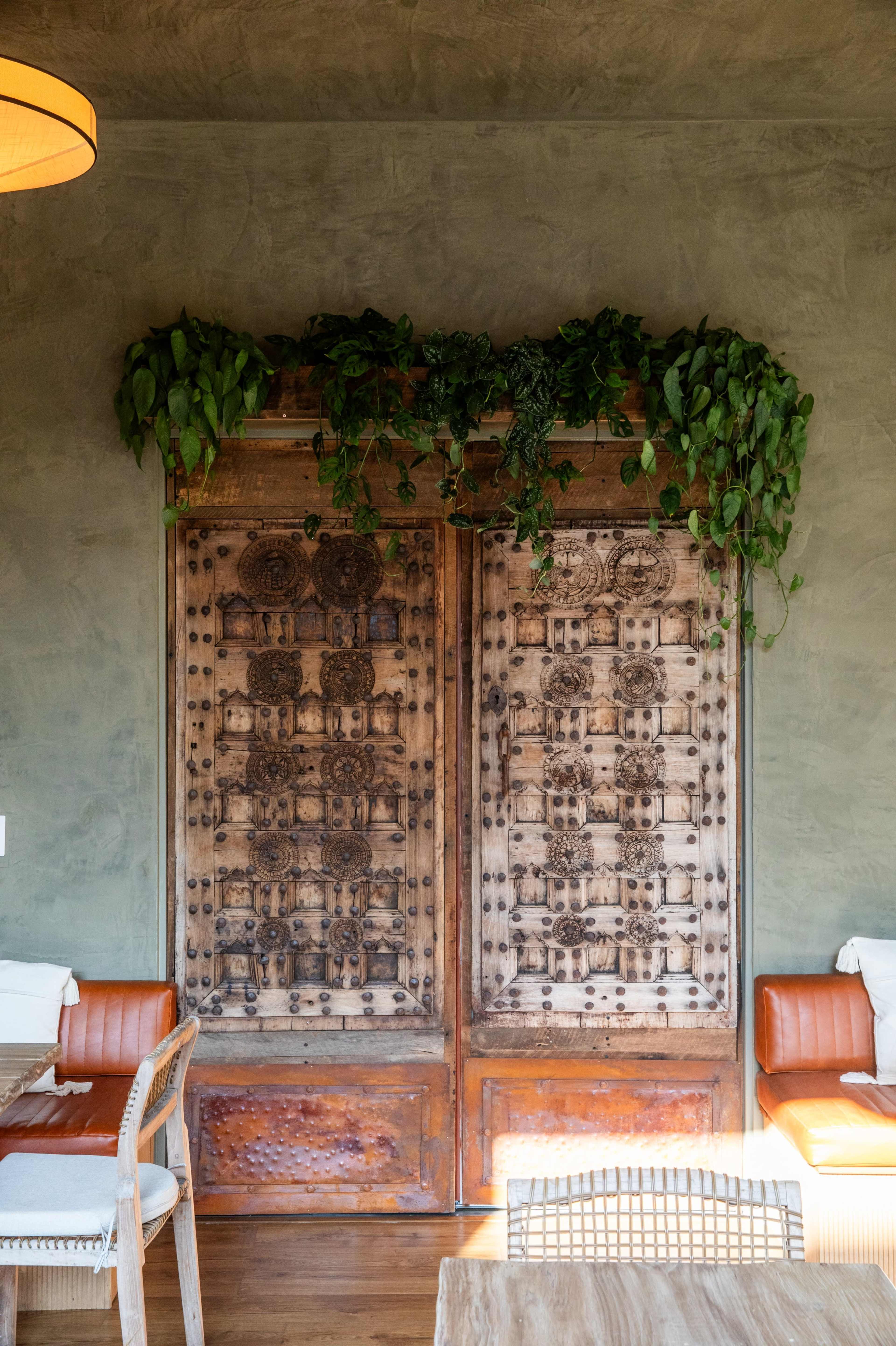 The image features a pair of intricately carved wooden doors adorned with greenery, positioned against a textured green wall in a restaurant setting.