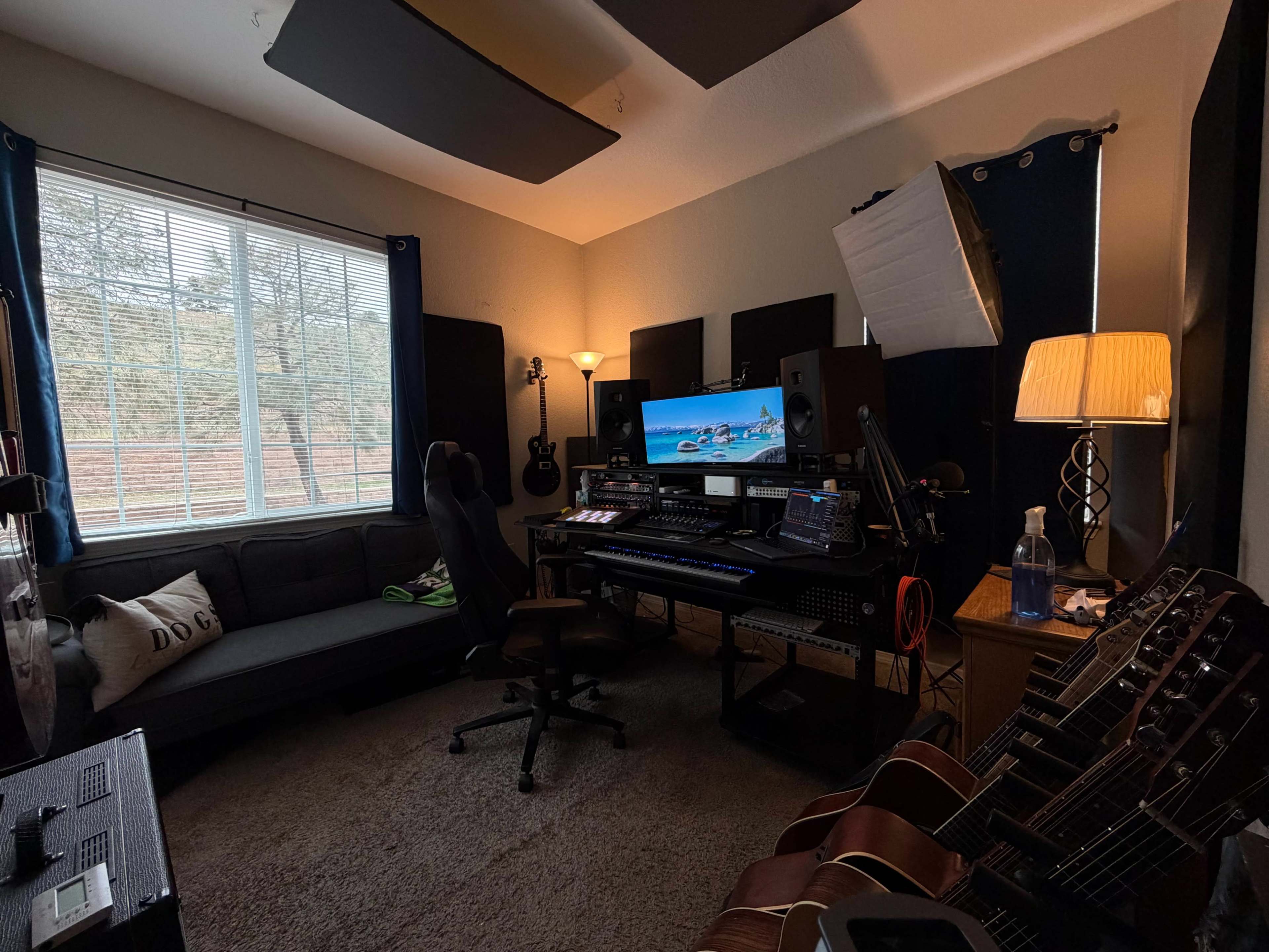 The image shows a home studio setup with a desk featuring musical equipment, a computer monitor, and several guitars positioned nearby.