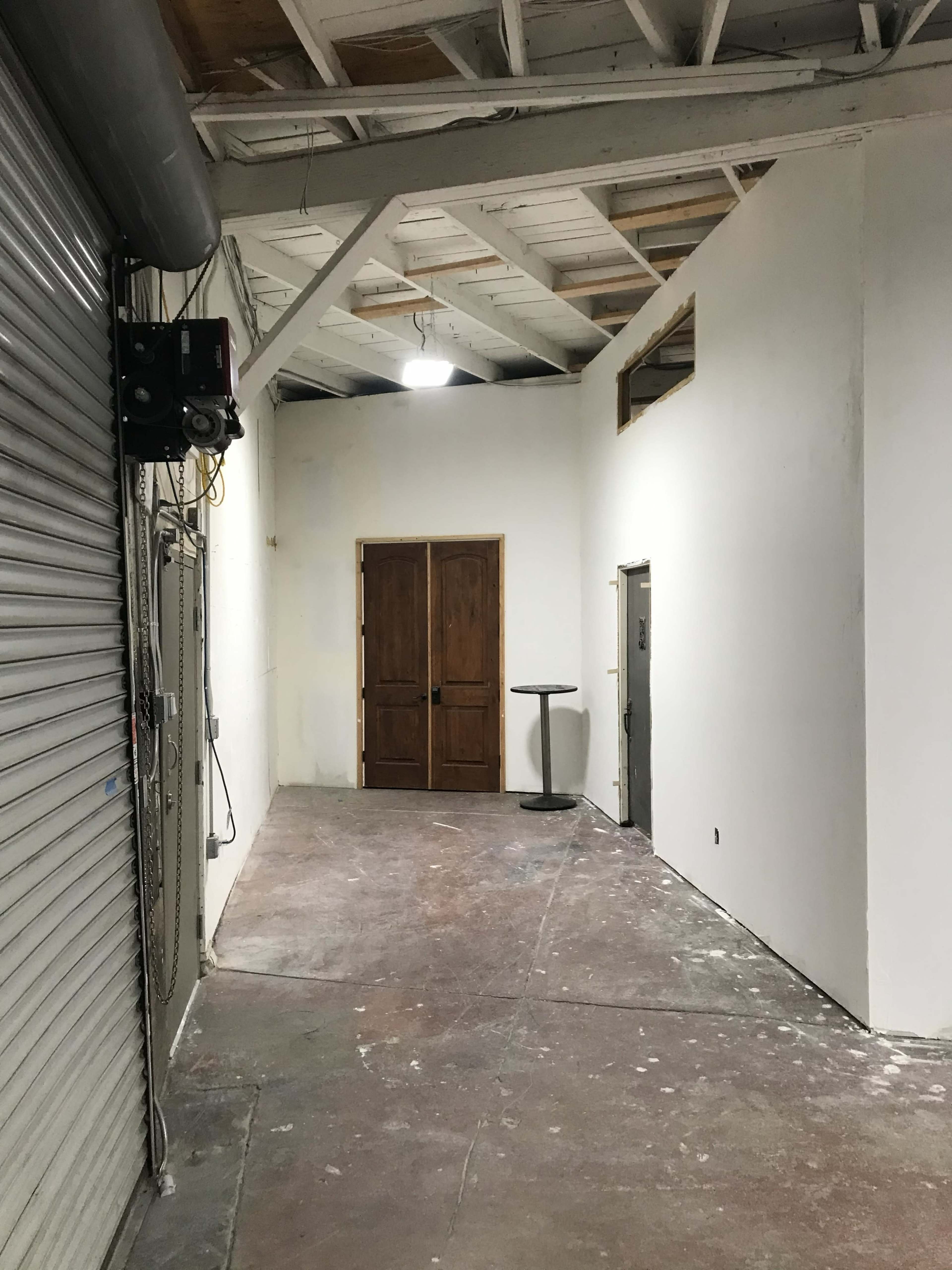 The image shows an empty corridor with a closed metal rolling door on the left, a pair of wooden doors at the end, and a small round table against the wall.