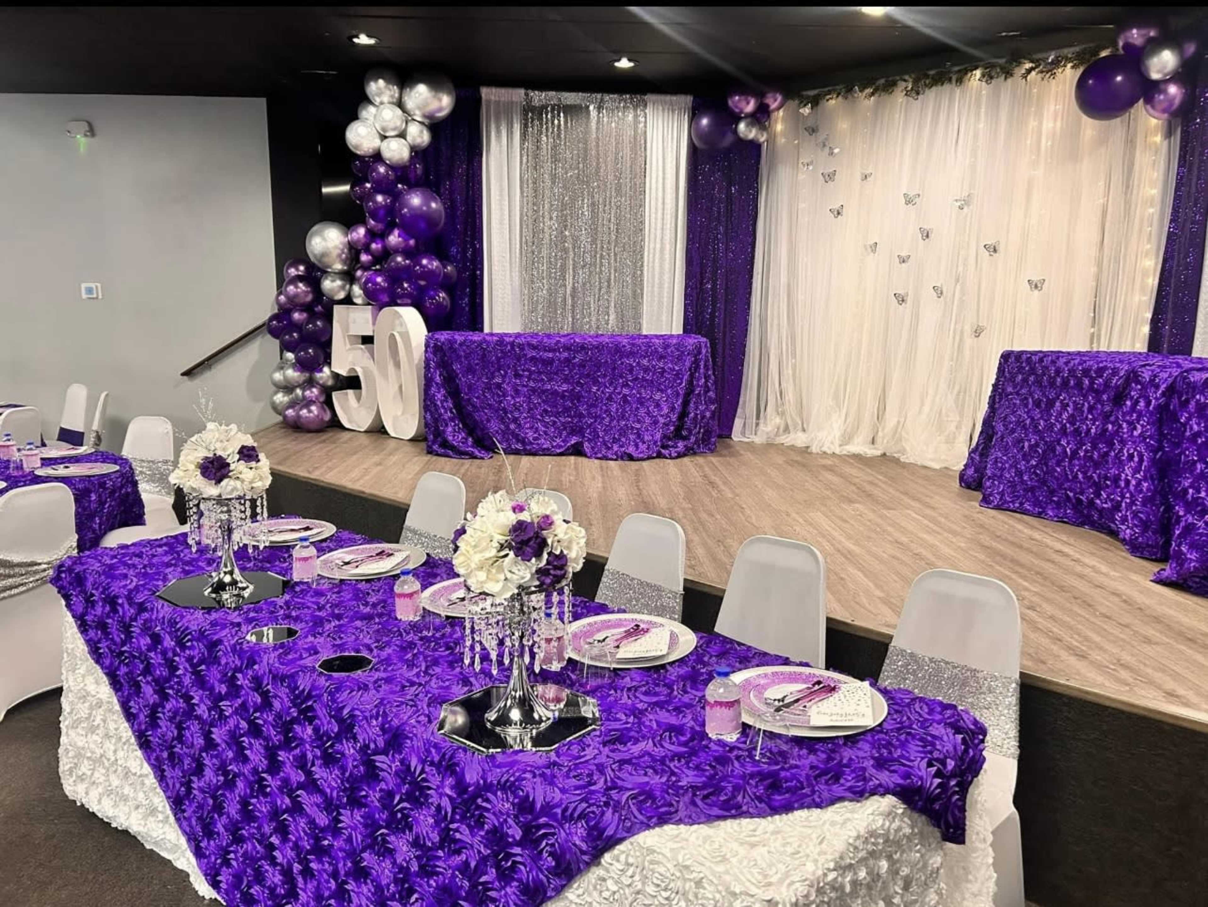 The image shows a decorated event space with purple tablecloths, silver accents, and a backdrop of draped fabric and balloons, designed for a 50th celebration.