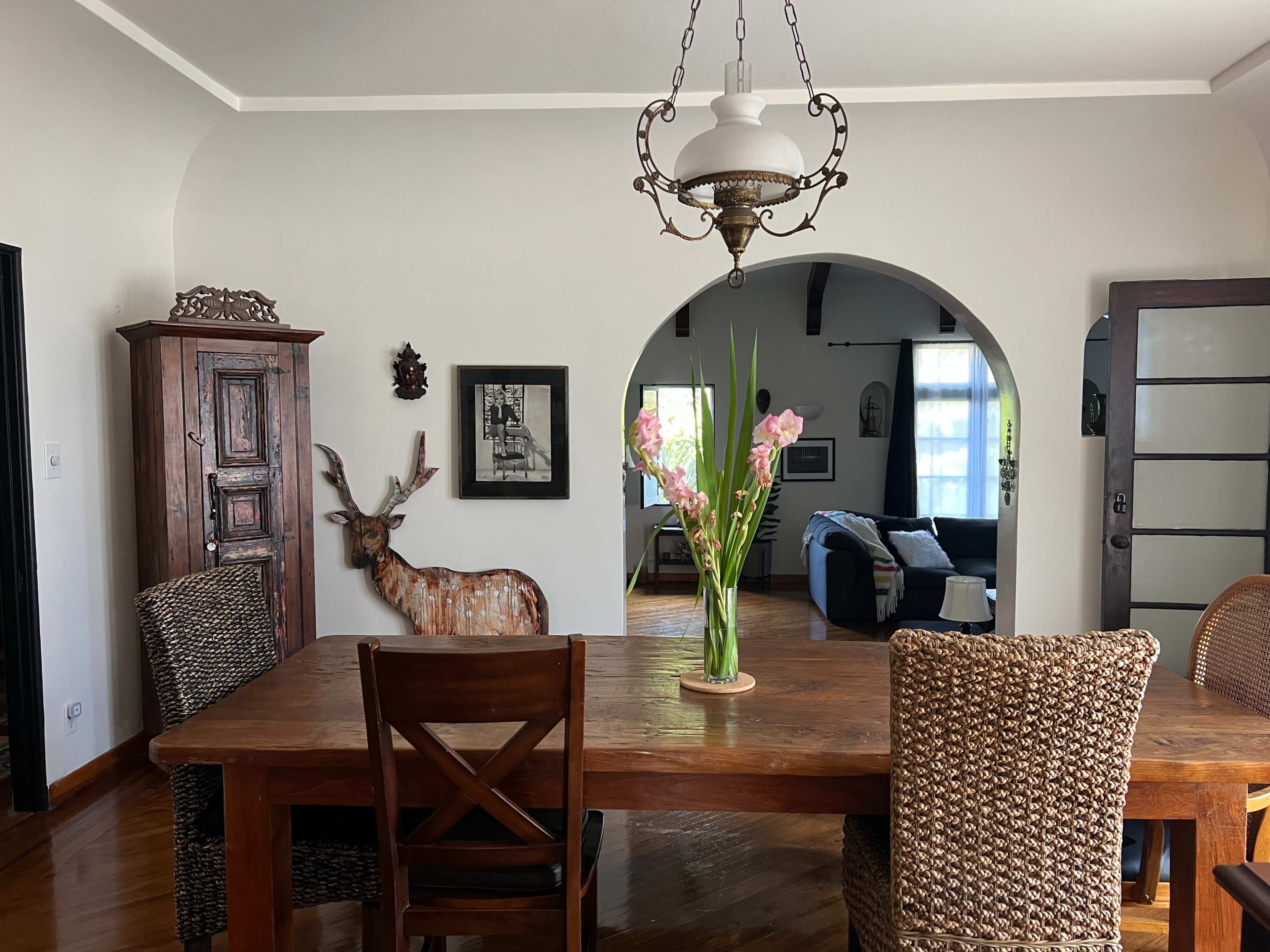 A wooden dining table with chairs is set in a room featuring an arched doorway, a deer sculpture, and a vase of flowers.