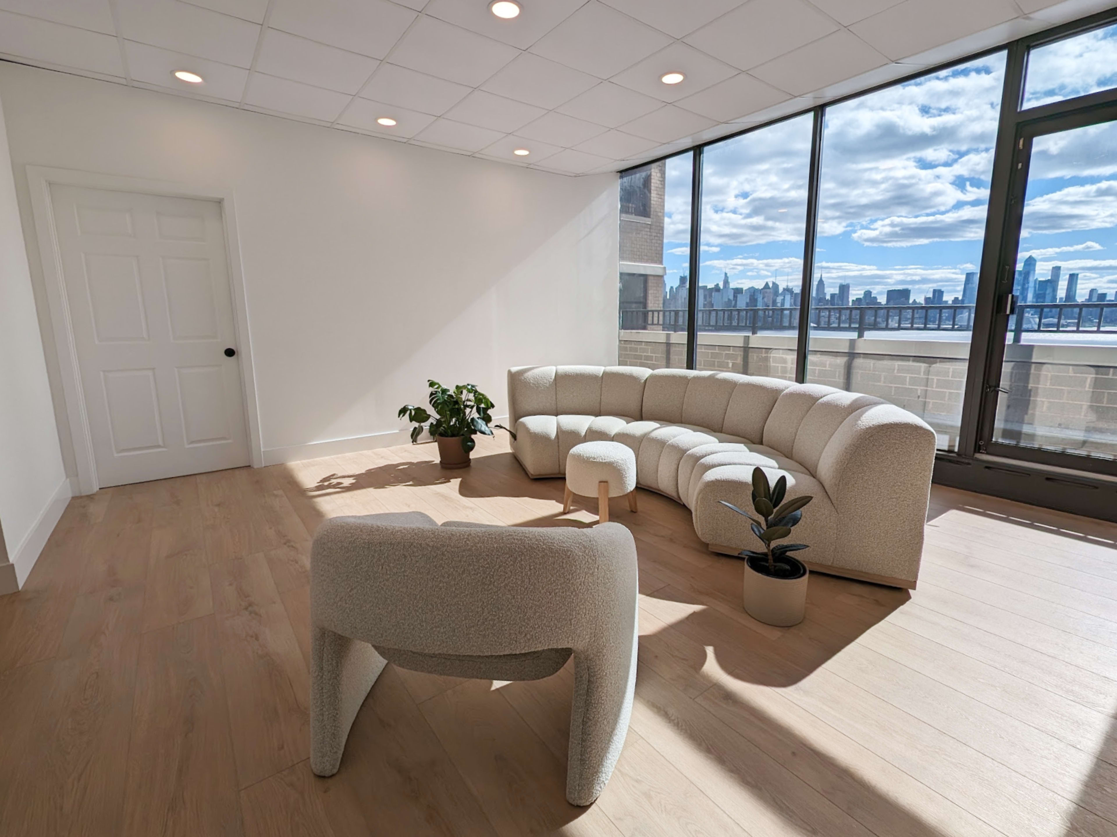 A modern, minimalist lounge area features a curved, light-colored sofa and a matching chair, with potted plants and large windows showcasing a city skyline.