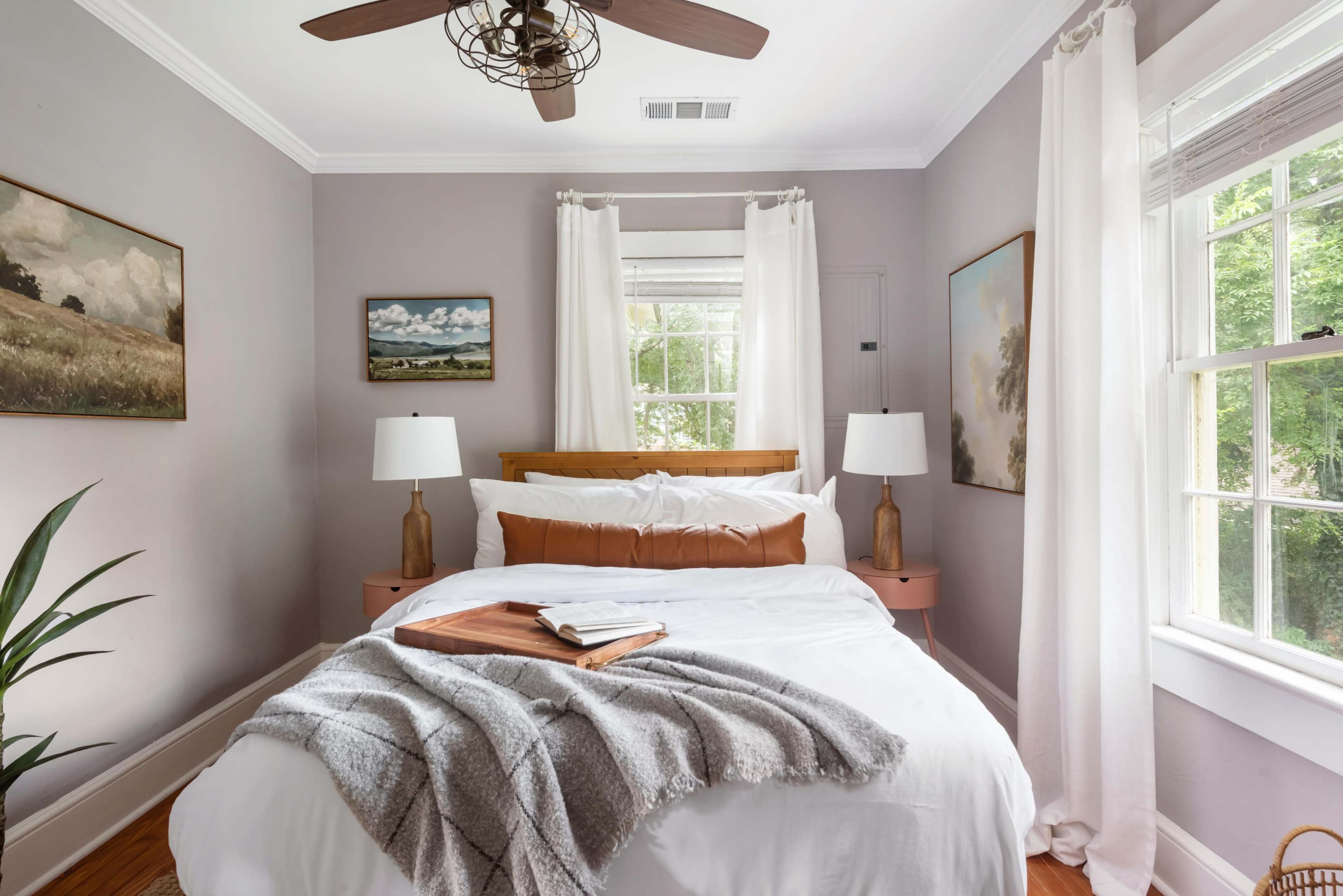 The image shows a cozy bedroom with a bed, two bedside lamps, artwork on the walls, and large windows partially covered by white curtains.