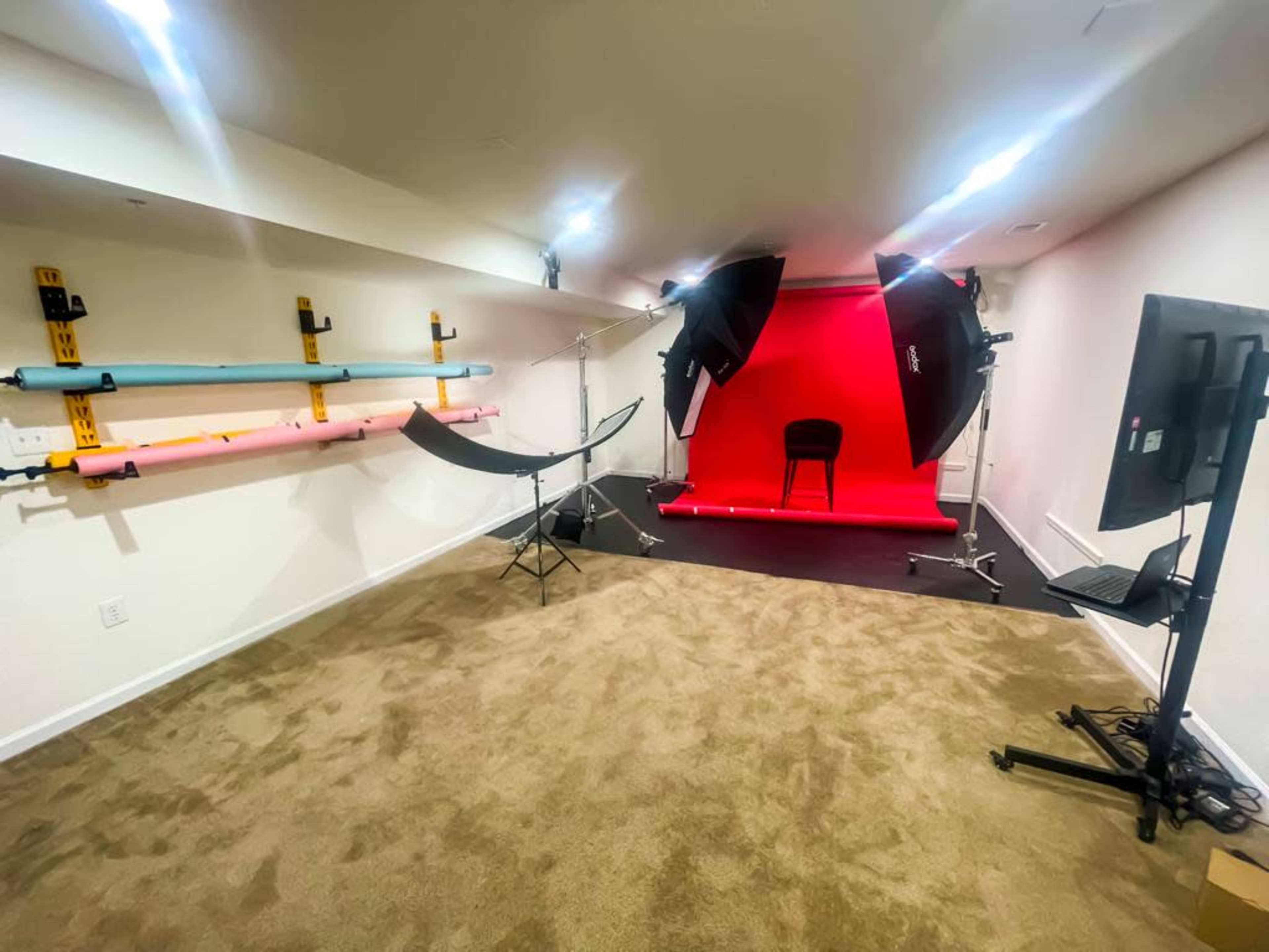 A photography studio featuring a red backdrop, studio lights, a chair, and various colored backdrop rolls on the wall.