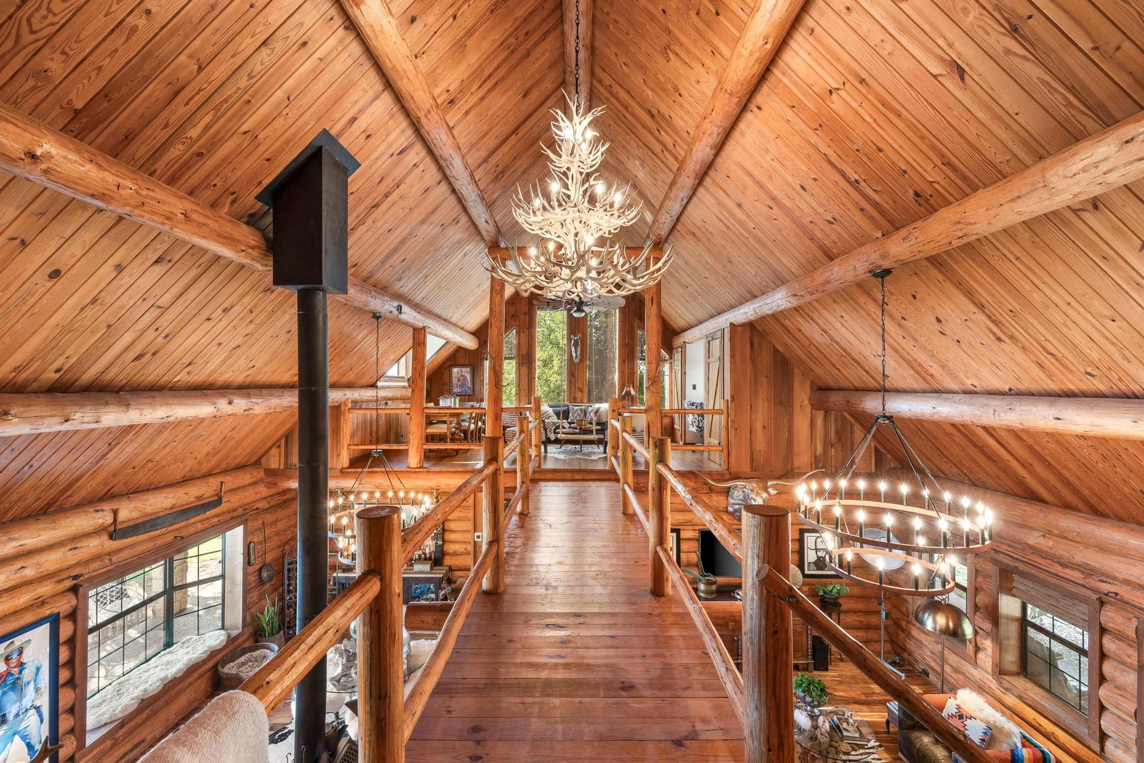 The image shows an interior view of a wooden cabin with high ceilings, exposed beams, and a balcony overlooking a living area.