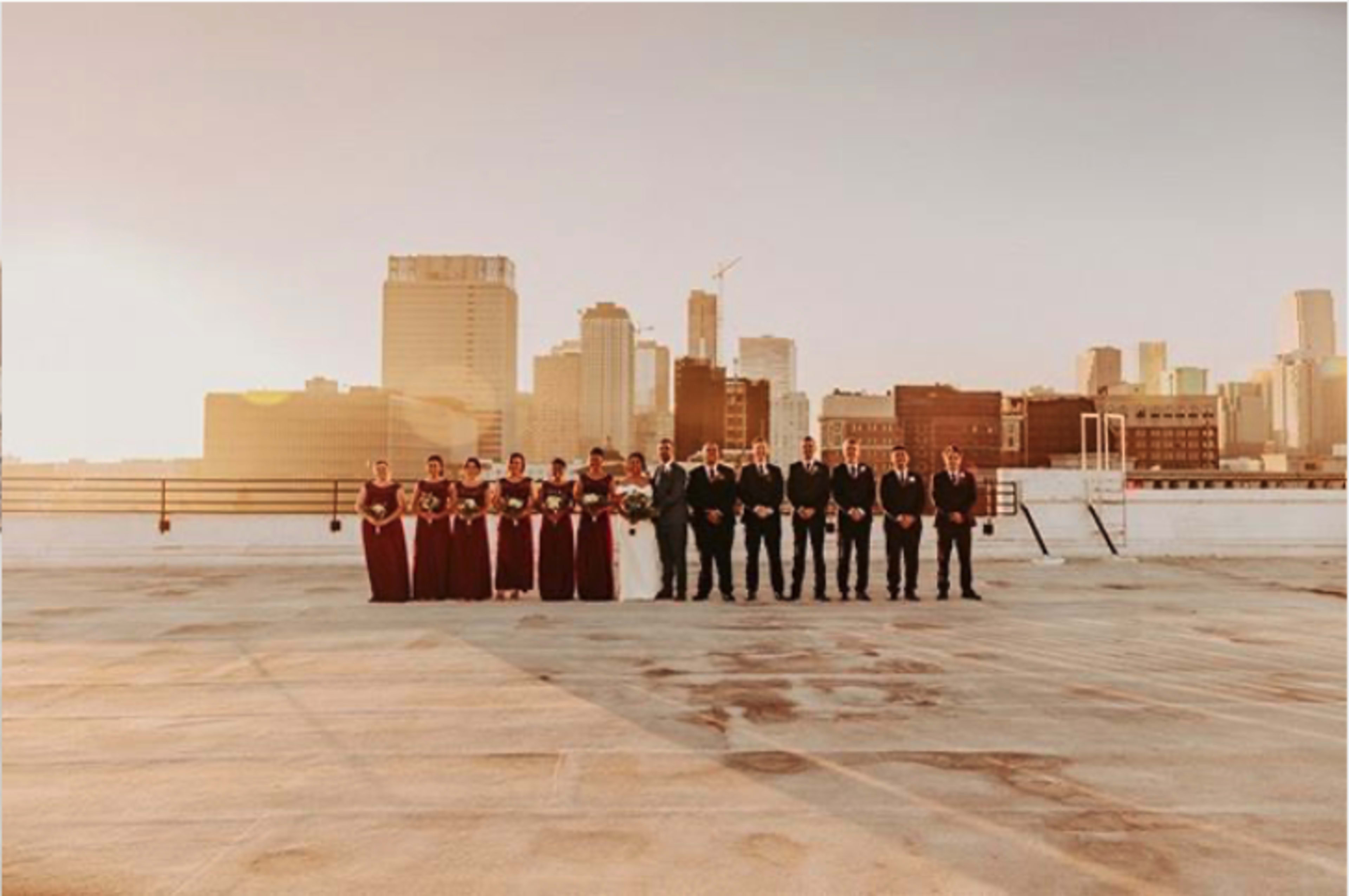 A wedding party in formal attire stands on a rooftop with a city skyline in the background during sunset.