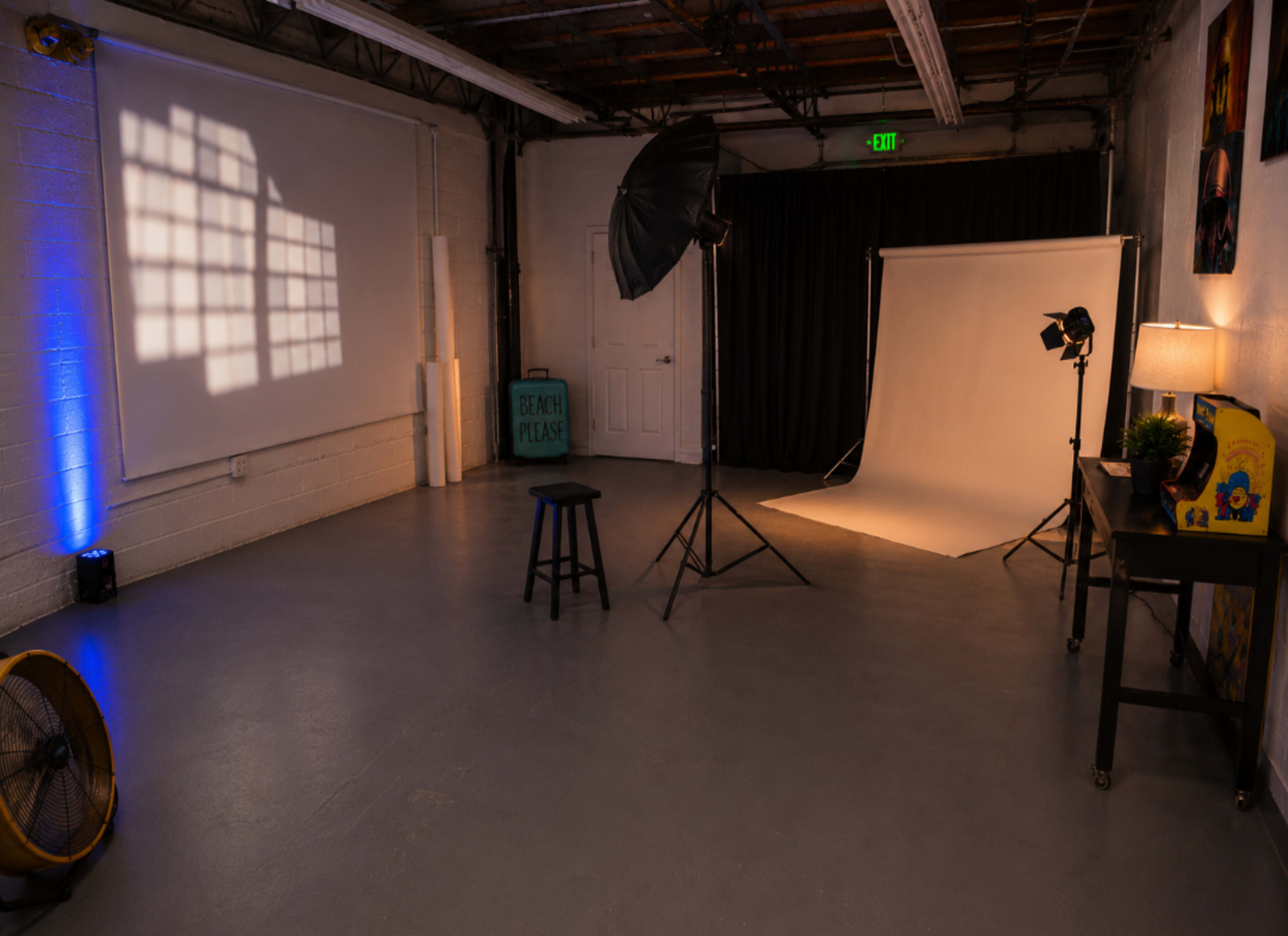 A photography studio features a backdrop, lighting equipment, a stool, and a fan in a spacious, well-lit area.