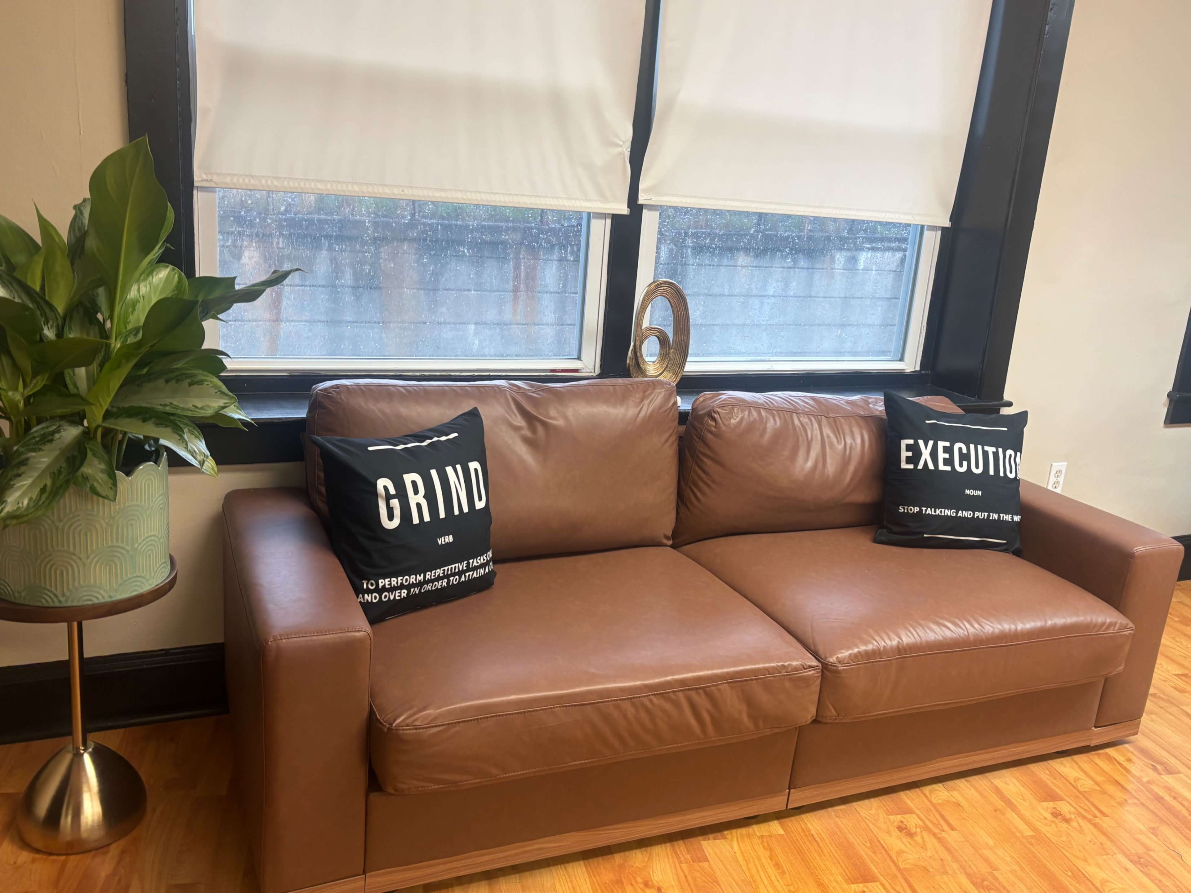The image shows a brown leather sofa with two black cushions that read "GRIND" and "EXECUTE," accompanied by a plant in a decorative pot and a side table.