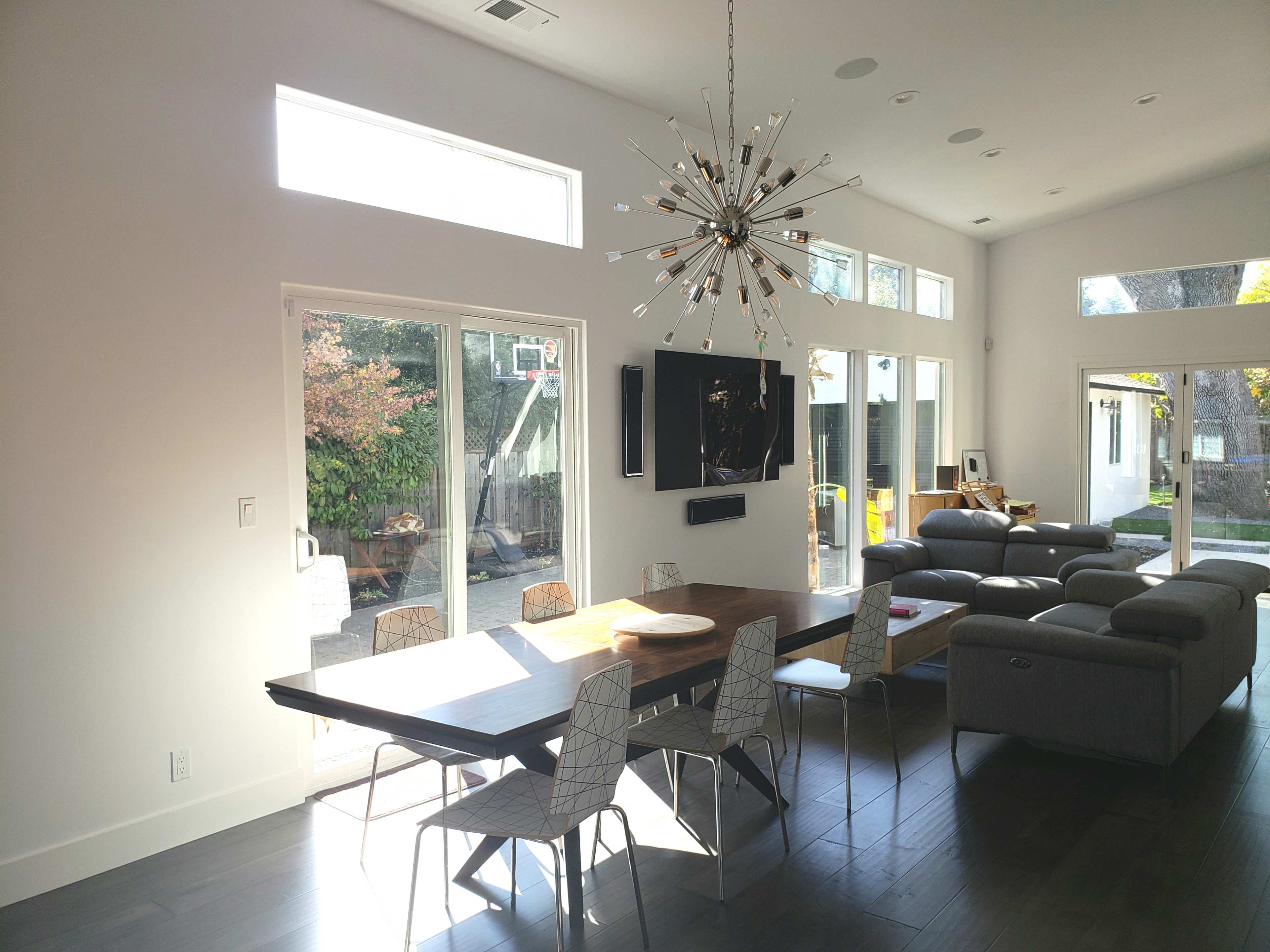 A modern dining area with a large wooden table, sleek chairs, and a starburst chandelier, adjacent to a living space with grey sofas and large windows.