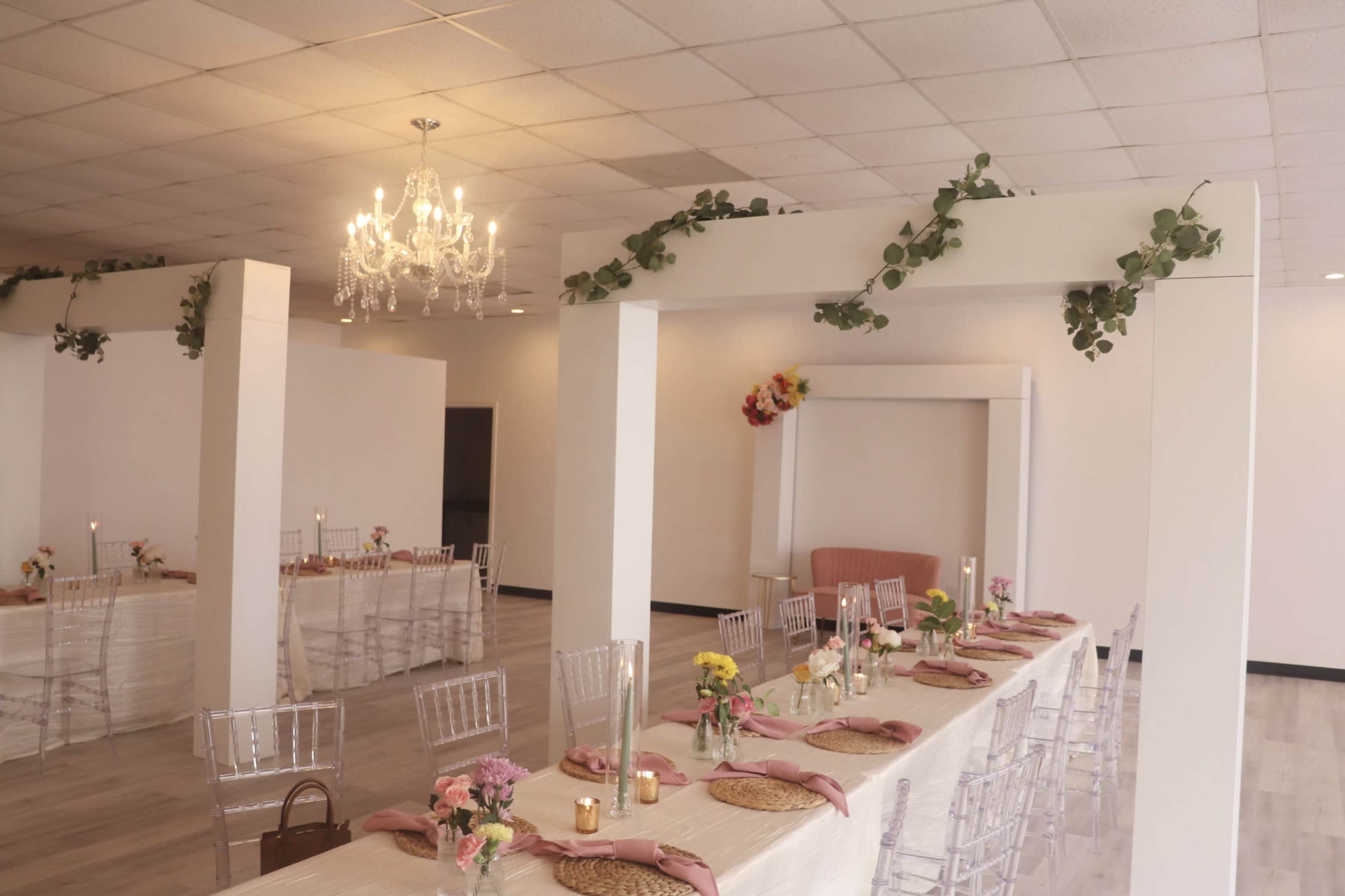 A spacious, elegantly decorated event venue features long tables adorned with floral centerpieces and soft lighting under a chandelier.