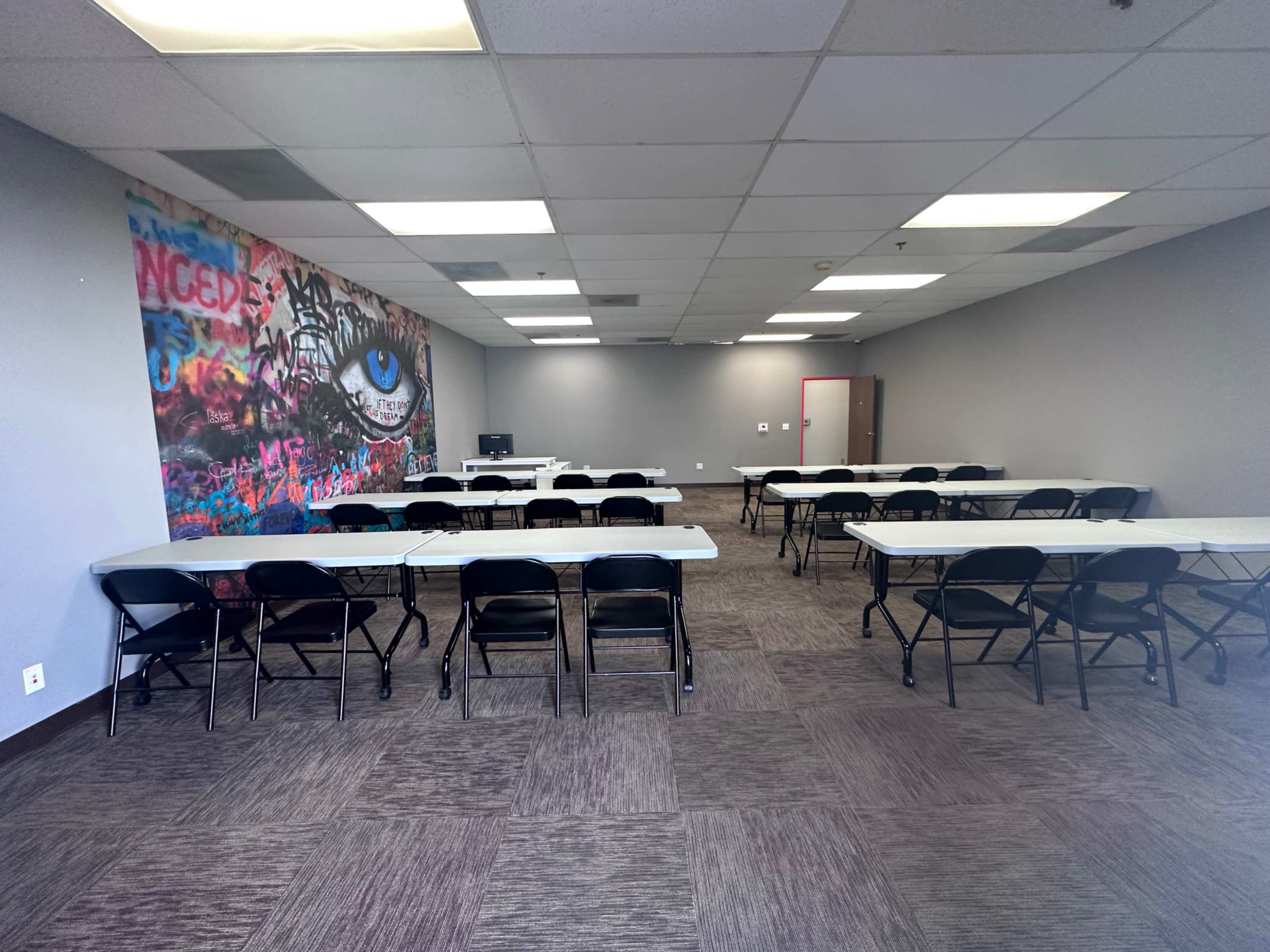 A spacious classroom with several rows of folding tables and chairs arranged neatly, alongside a colorful mural featuring an eye on one wall.