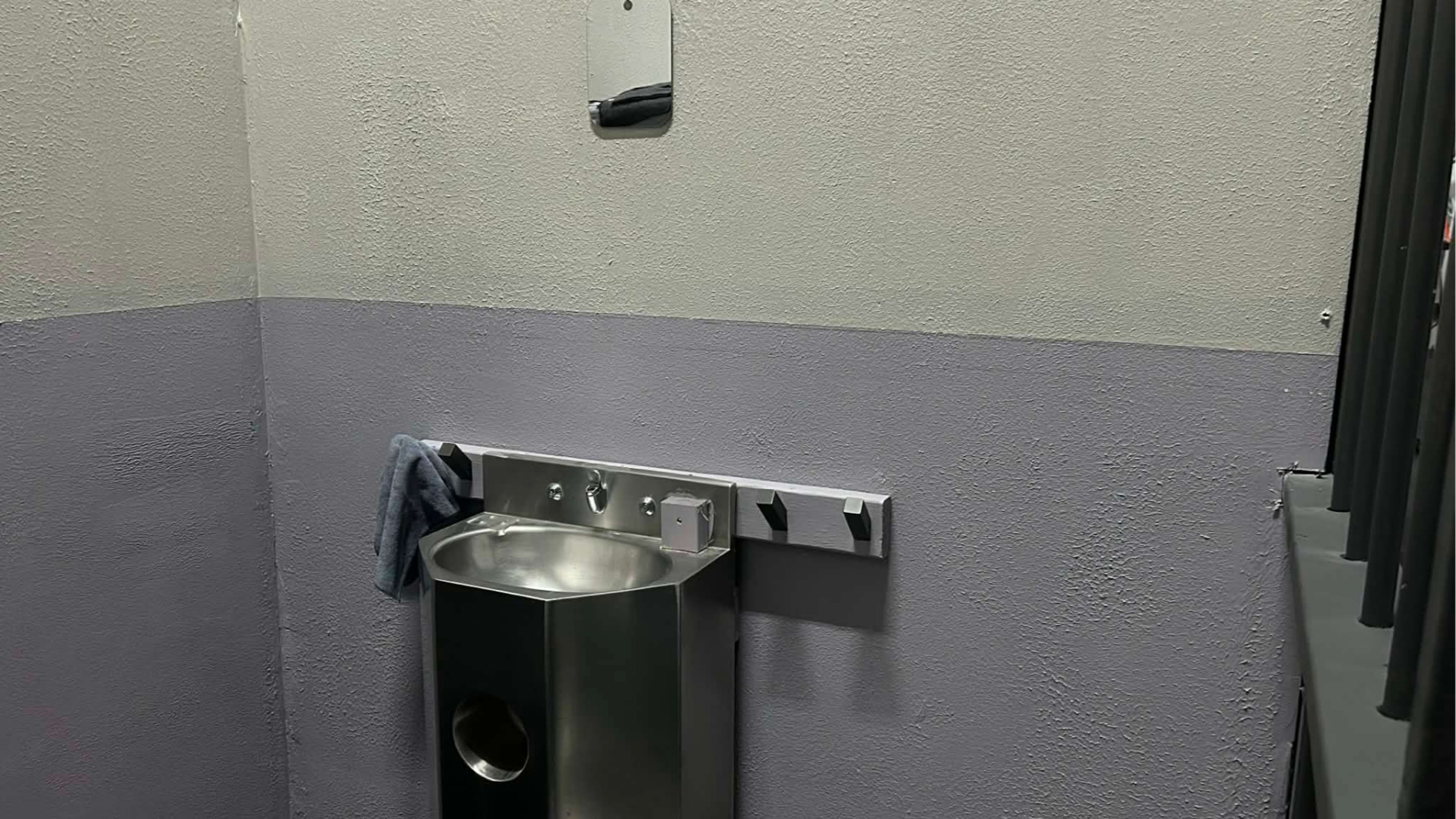 The image shows a stainless steel drinking fountain mounted on a wall, beside a gray and purple painted surface, with a towel hanging on a nearby hook.