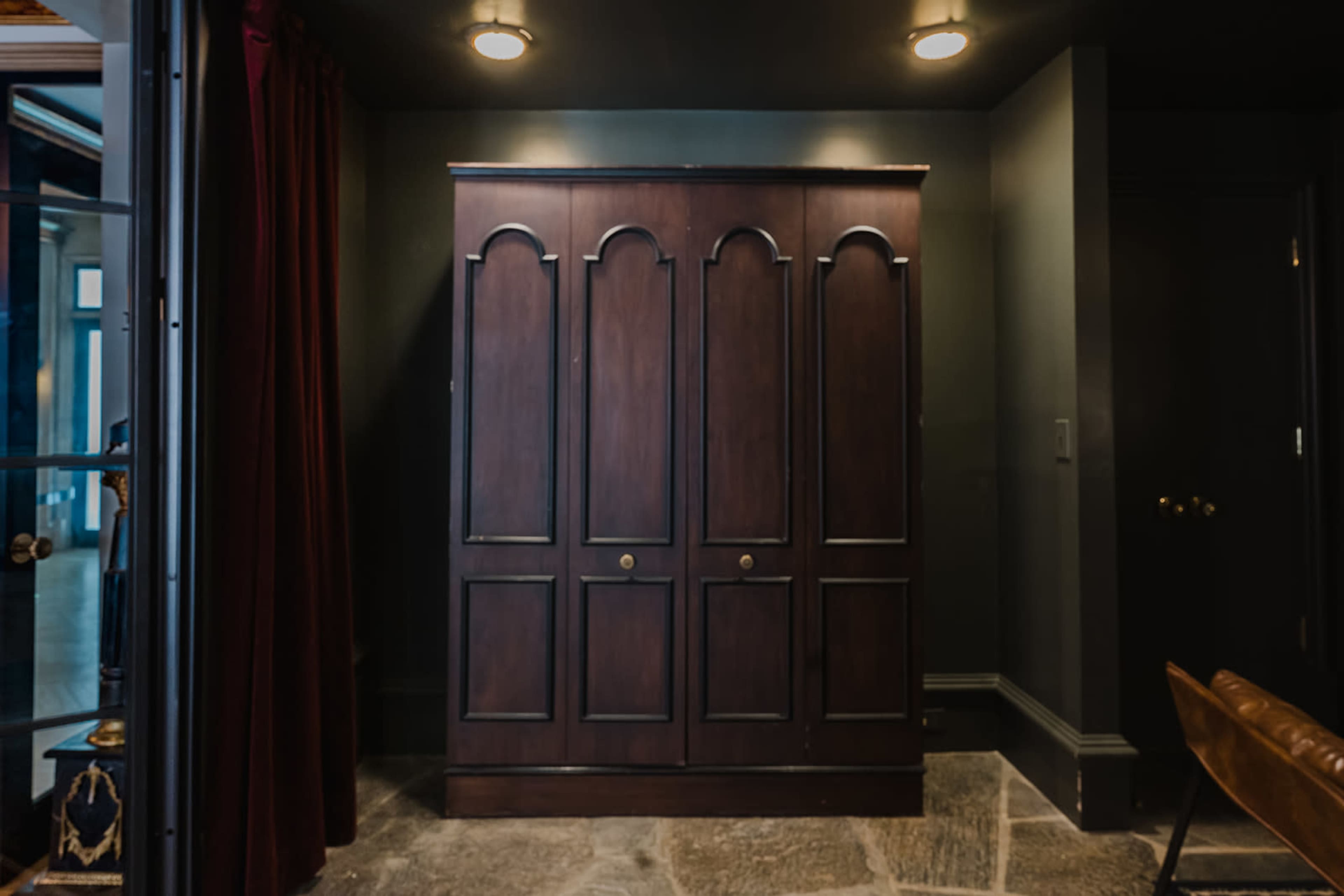 A dark wooden wardrobe with arched panels is positioned against a gray wall in a dimly lit room.