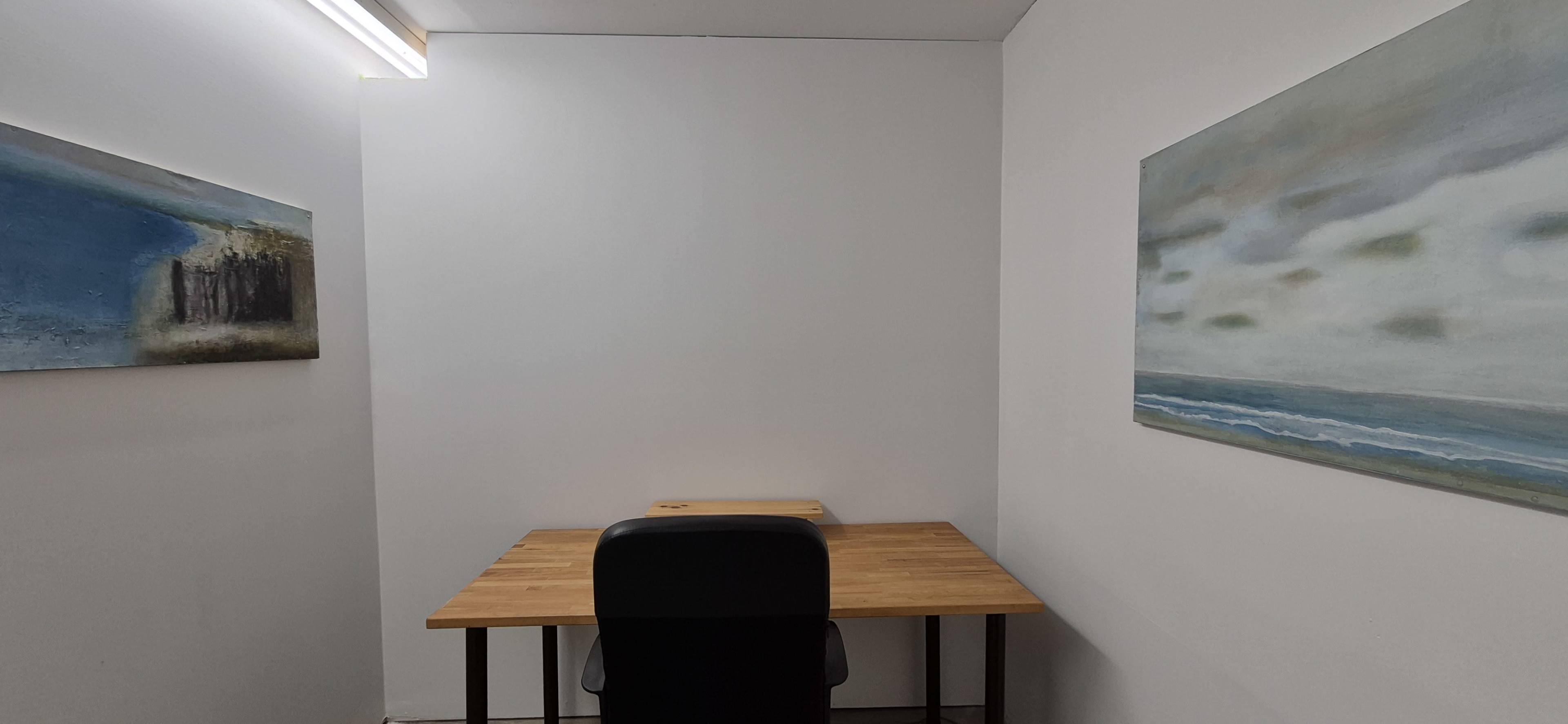 The image shows a minimalist office space with a wooden desk and a black chair, flanked by two paintings on the walls.