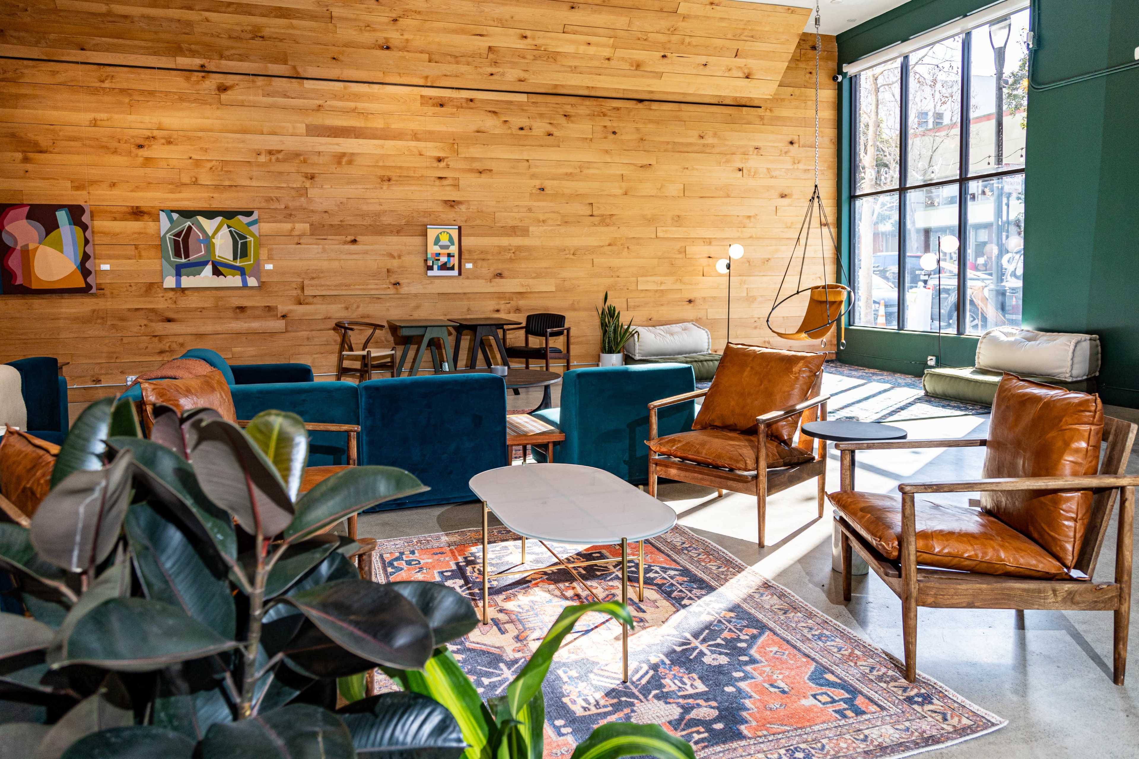 A modern lounge area features leather armchairs, a patterned rug, and a wooden accent wall, illuminated by natural light from large windows.