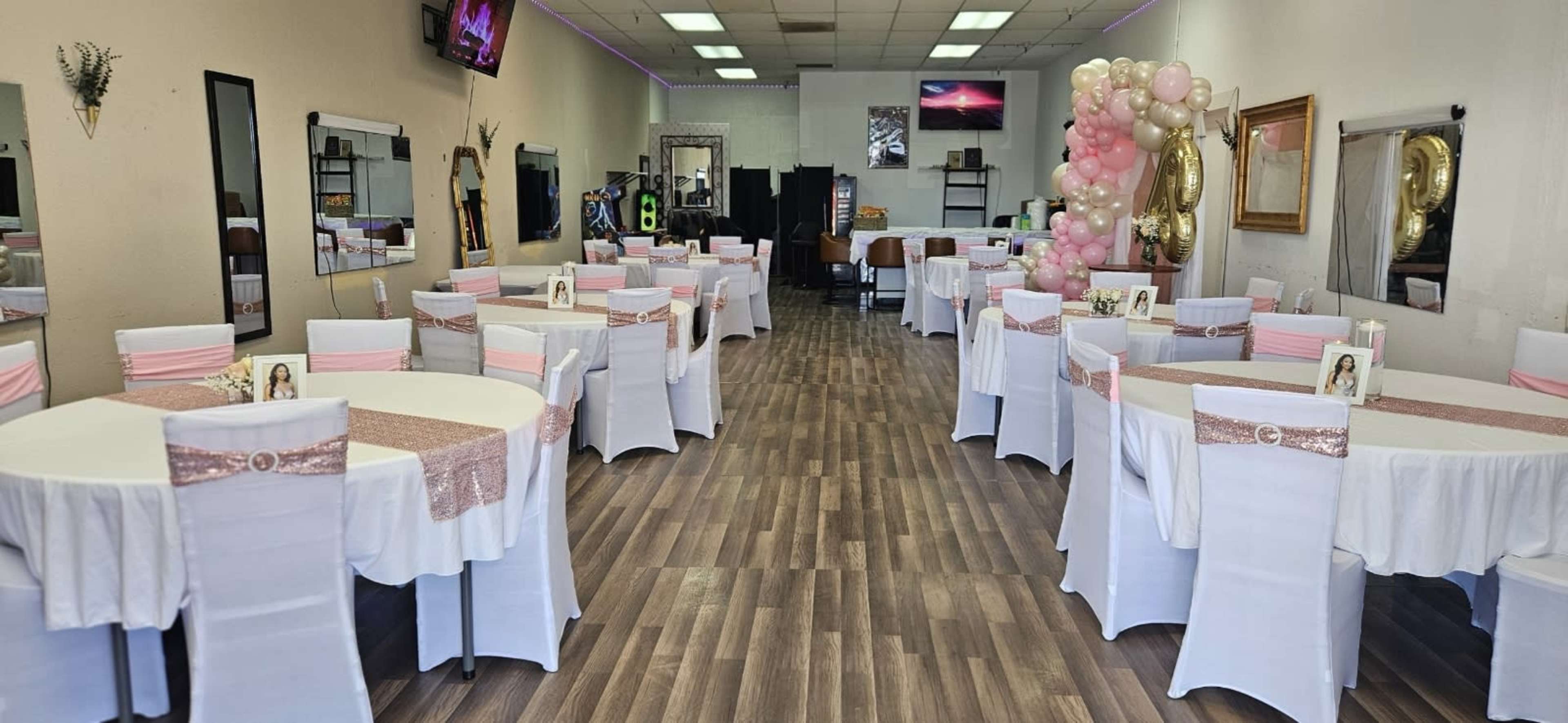 The image shows a decorated event space with several round tables covered in white tablecloths and pink accents, arranged neatly for a gathering or celebration.