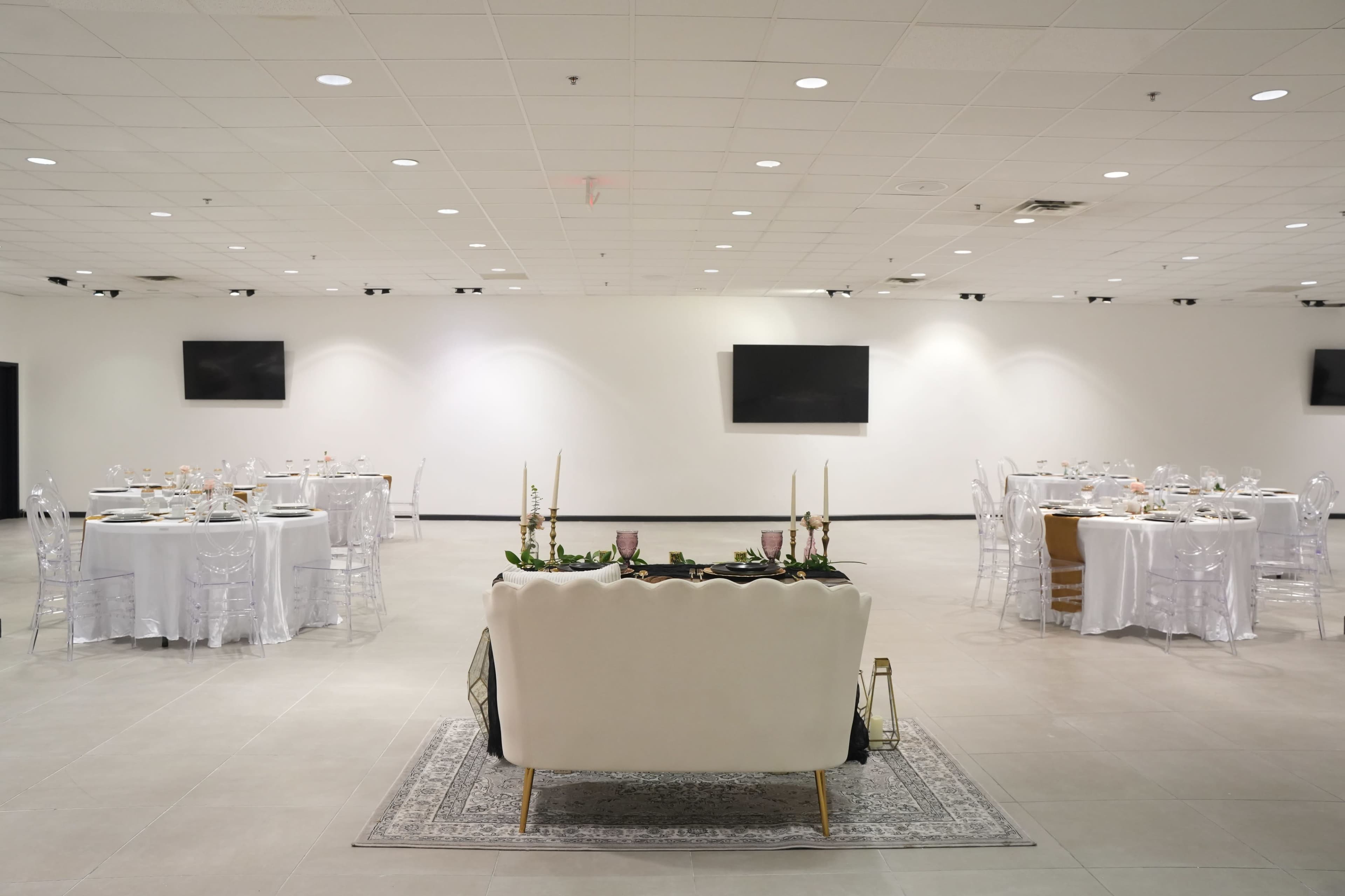 A spacious banquet hall features round tables set for dining and a white couch positioned centrally, with minimal decor and wall-mounted screens.