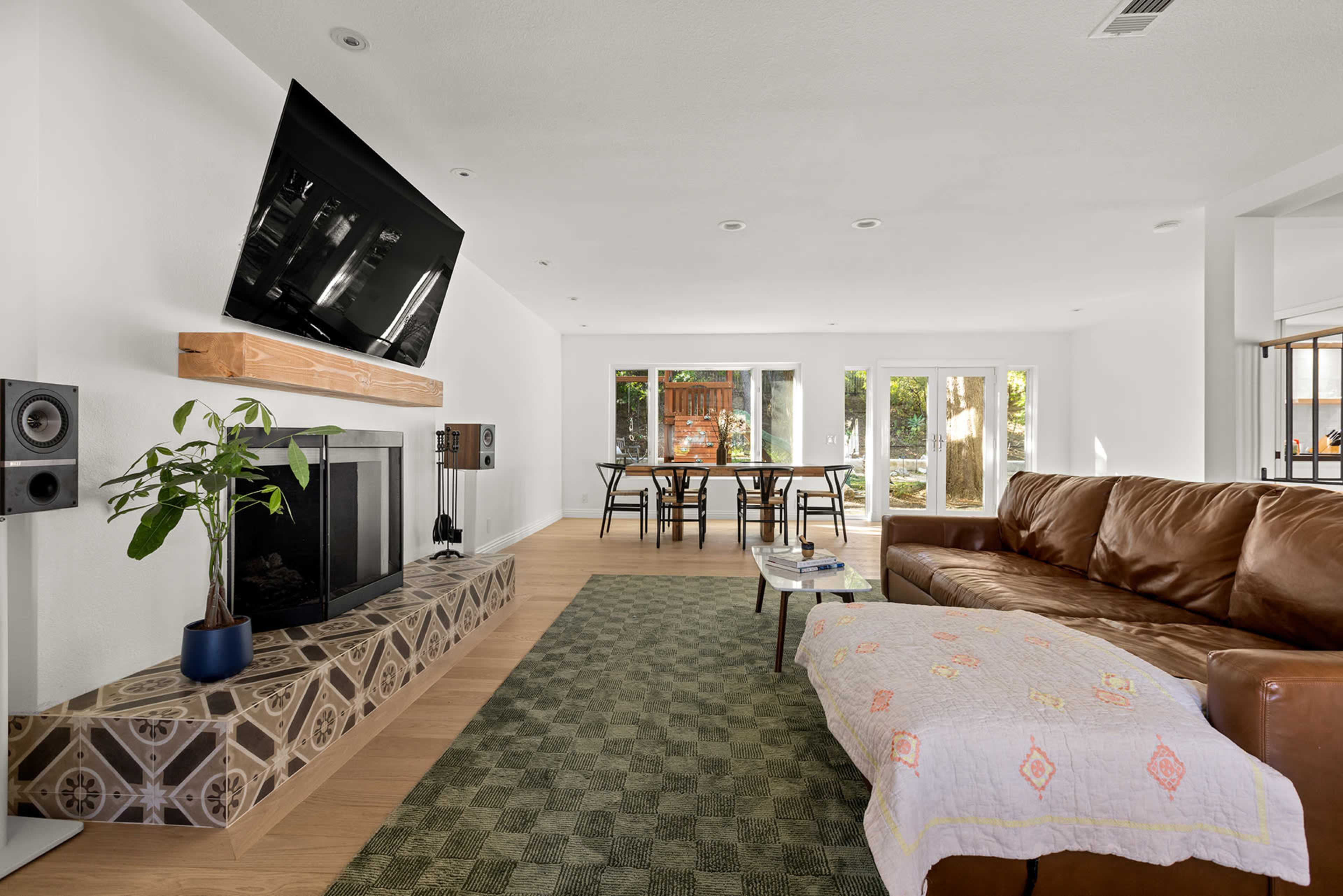 The living room features a leather sofa, a mounted television above a fireplace, and a dining table with chairs visible through large glass doors.