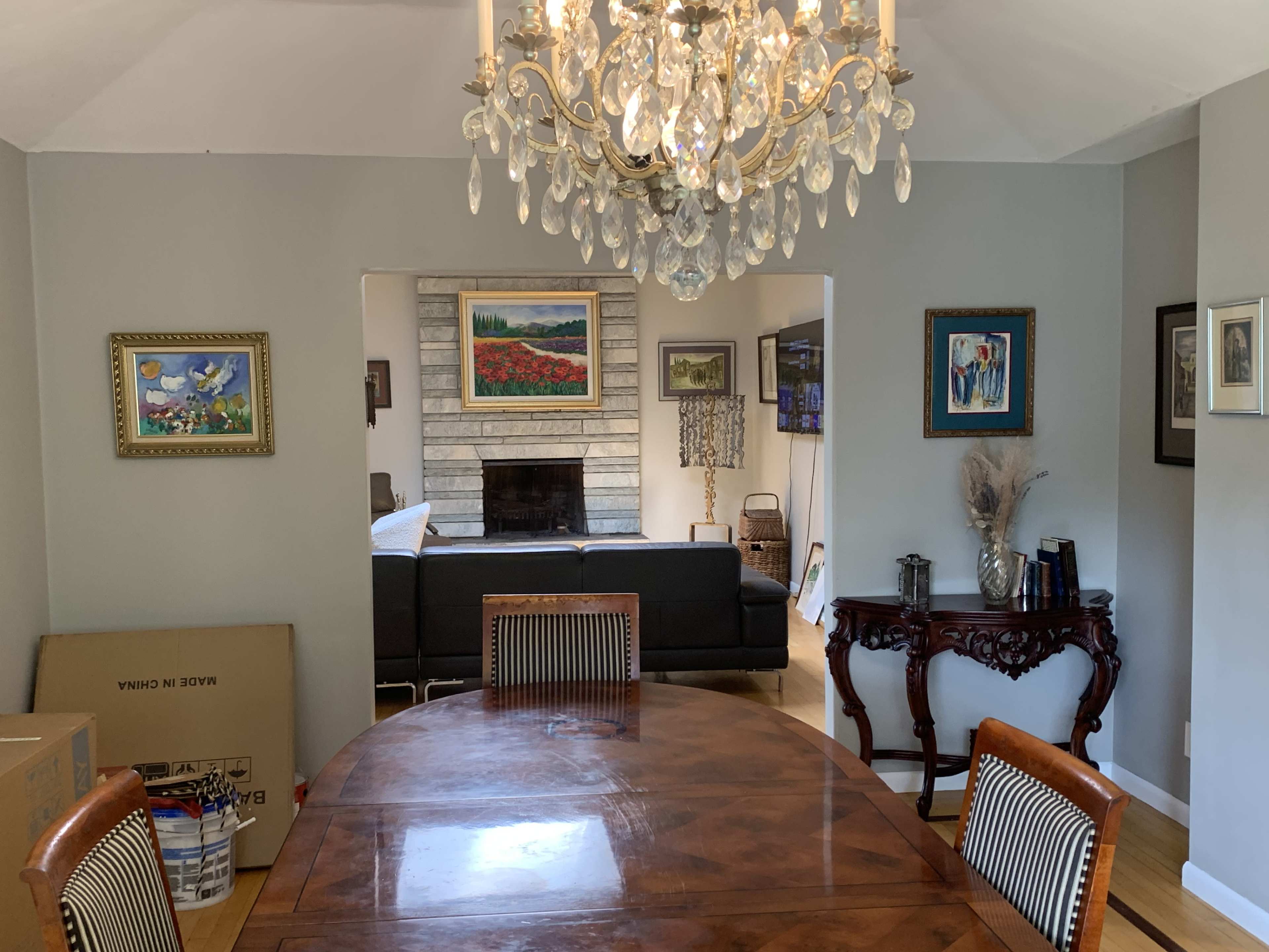 A dining room features a wooden table surrounded by chairs, with a chandelier overhead and a view into a living area with a stone fireplace.