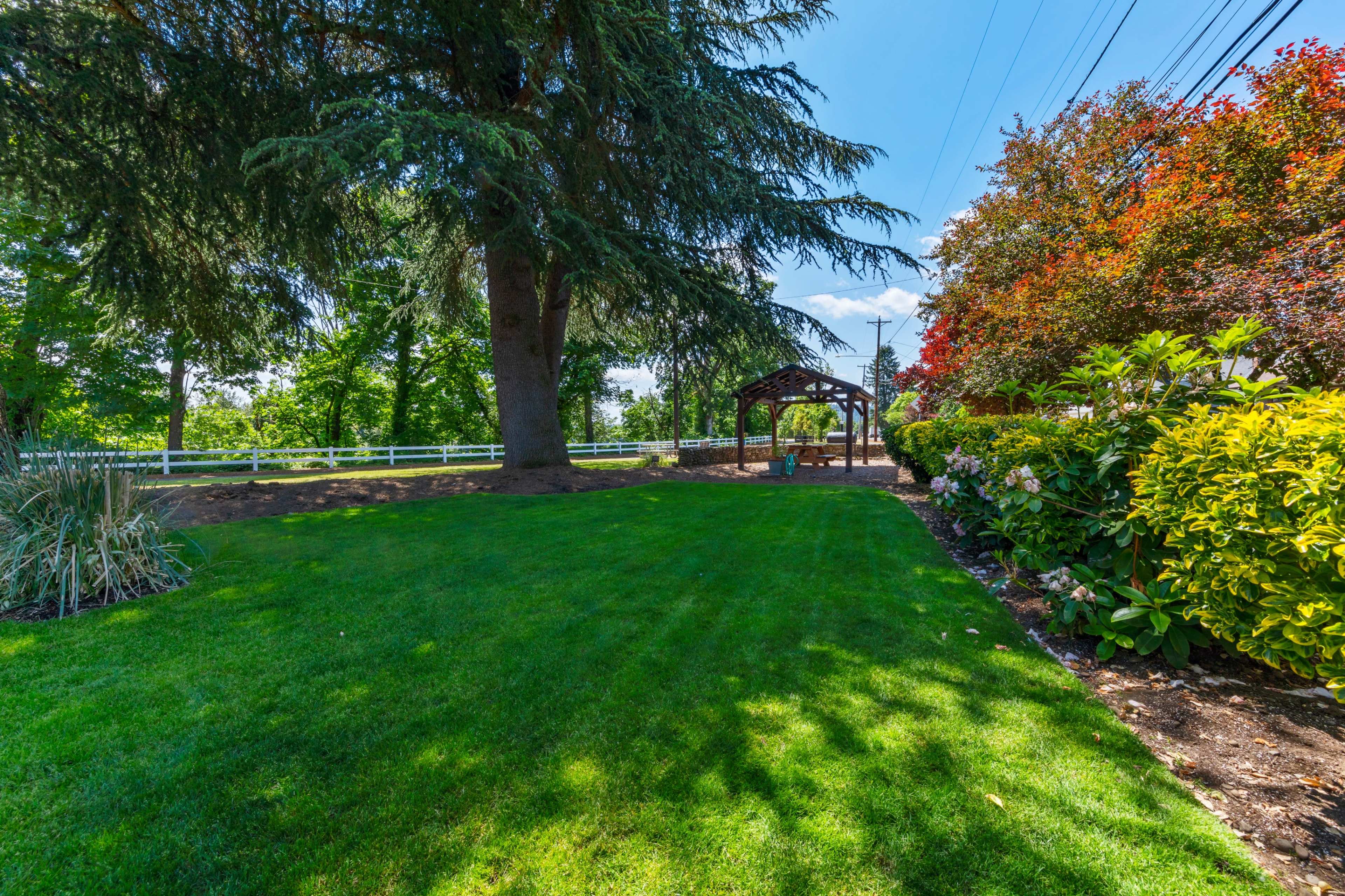 The image shows a landscaped garden with neatly trimmed grass, a wooden gazebo, and colorful flowering bushes under a clear blue sky.