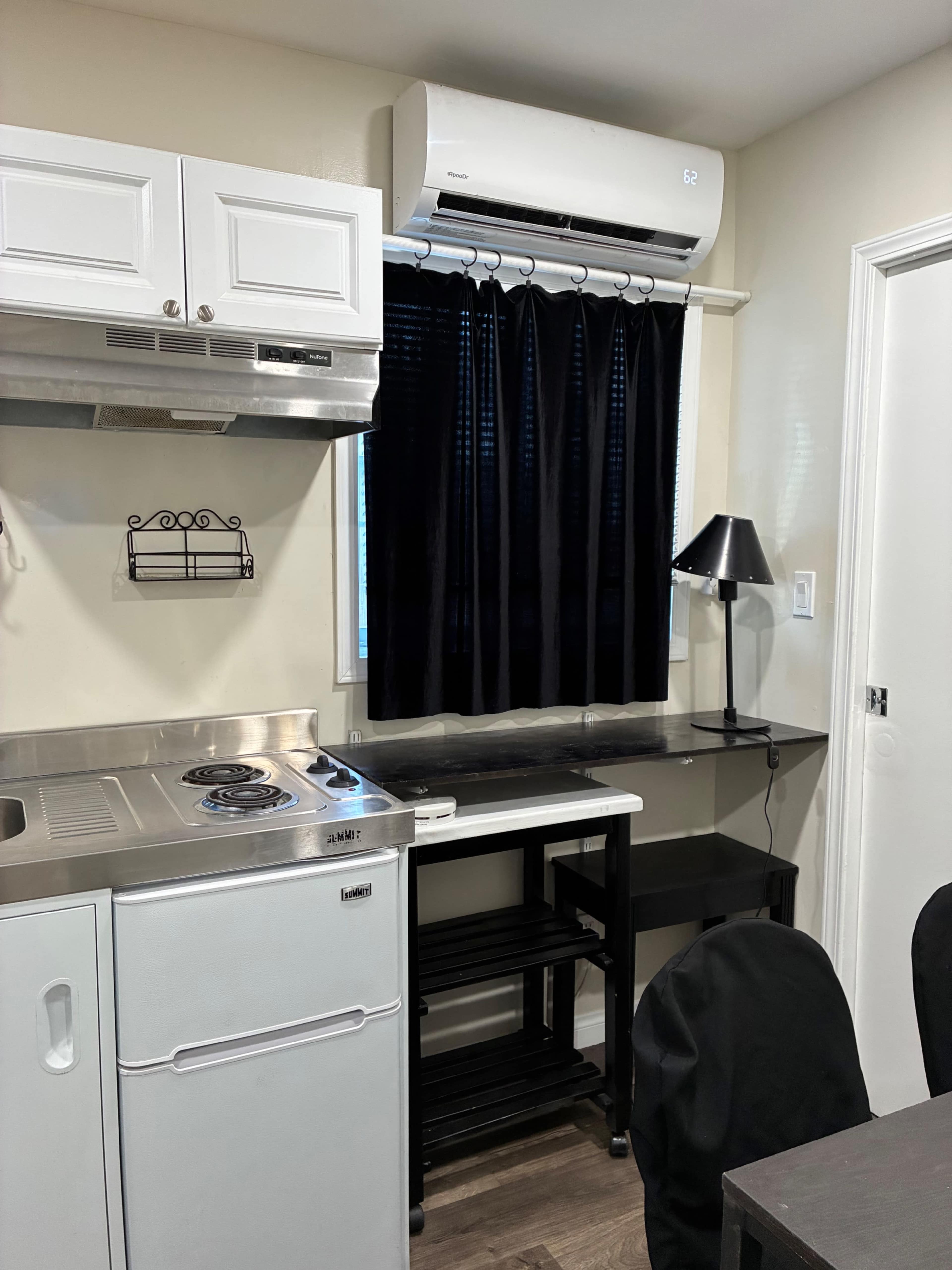 The image shows a compact kitchen area featuring a stove, sink, a small workspace with a lamp, and an air conditioning unit mounted above a window with black curtains.