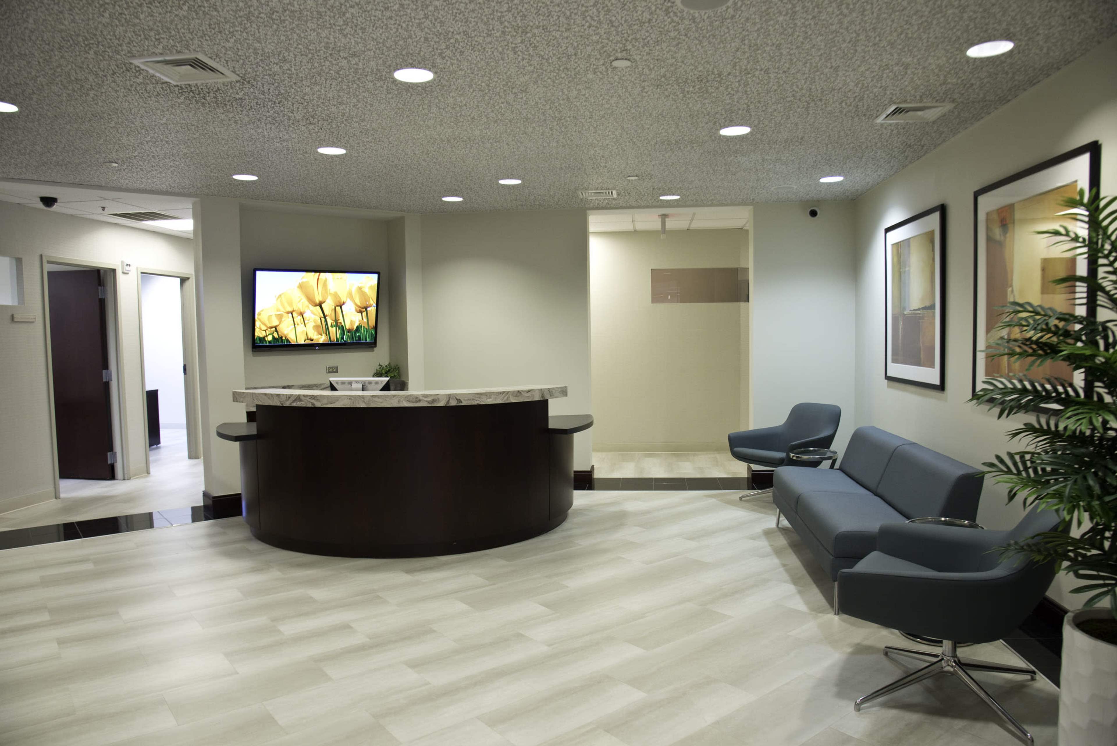 The image shows a modern reception area with a curved desk, a television displaying artwork, a gray seating area, and potted plants.