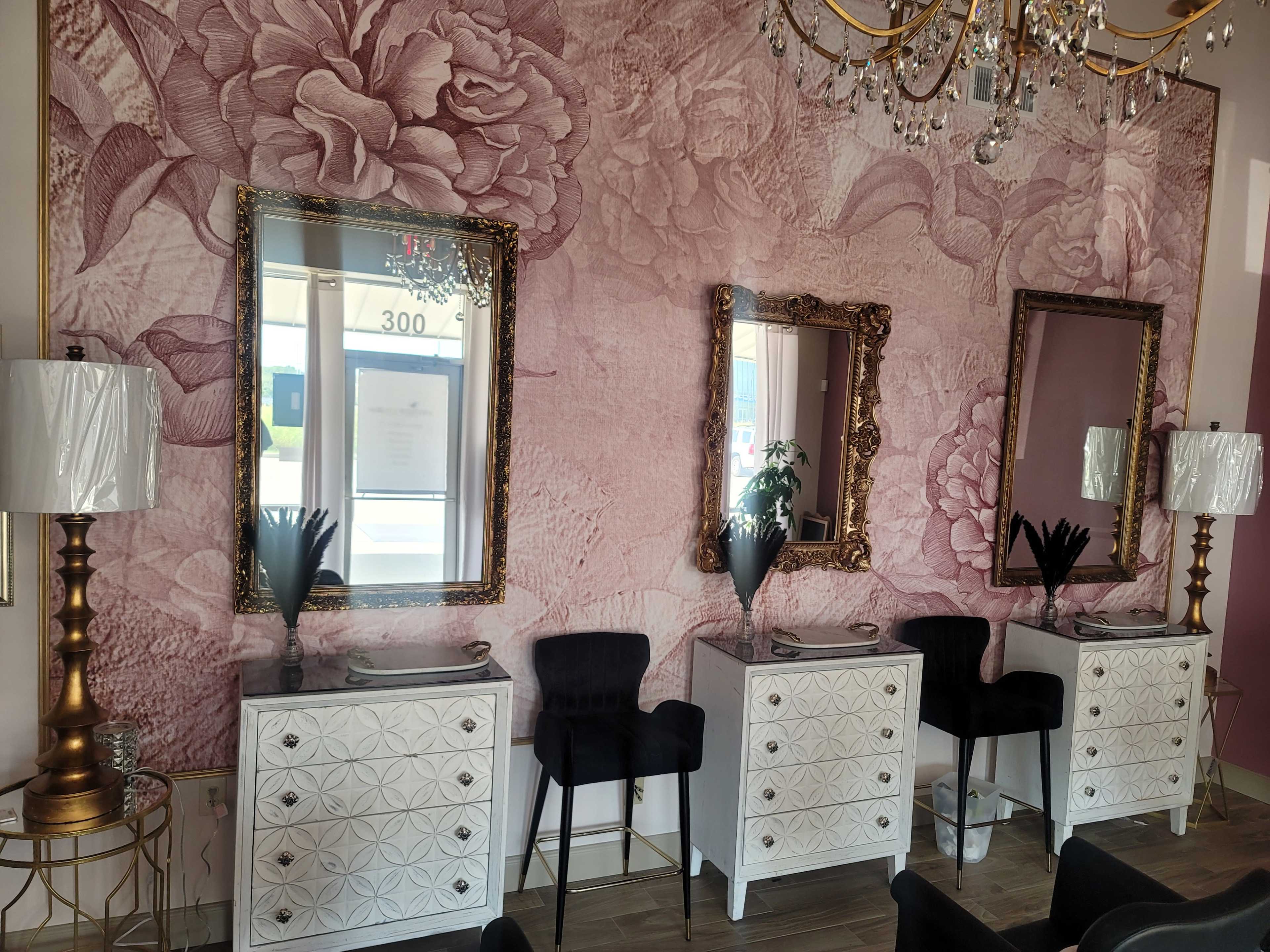 A stylish interior featuring three ornate mirrors above white dressers, flanked by black chairs and a lamp on a gold stand, all against a floral-patterned pink wallpaper.