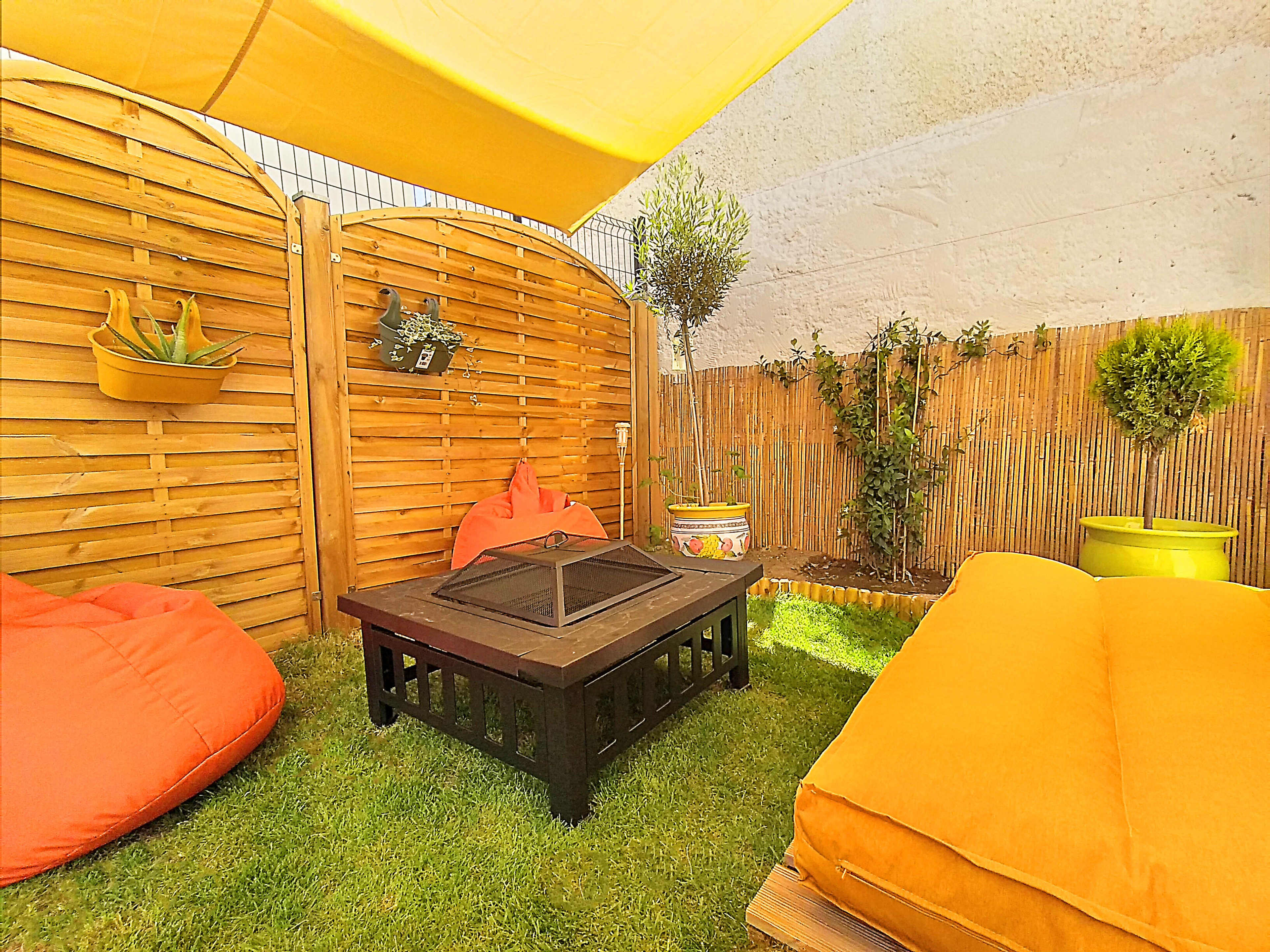 A cozy outdoor seating area with a black coffee table, bright orange cushions, and wooden privacy screens surrounded by greenery.