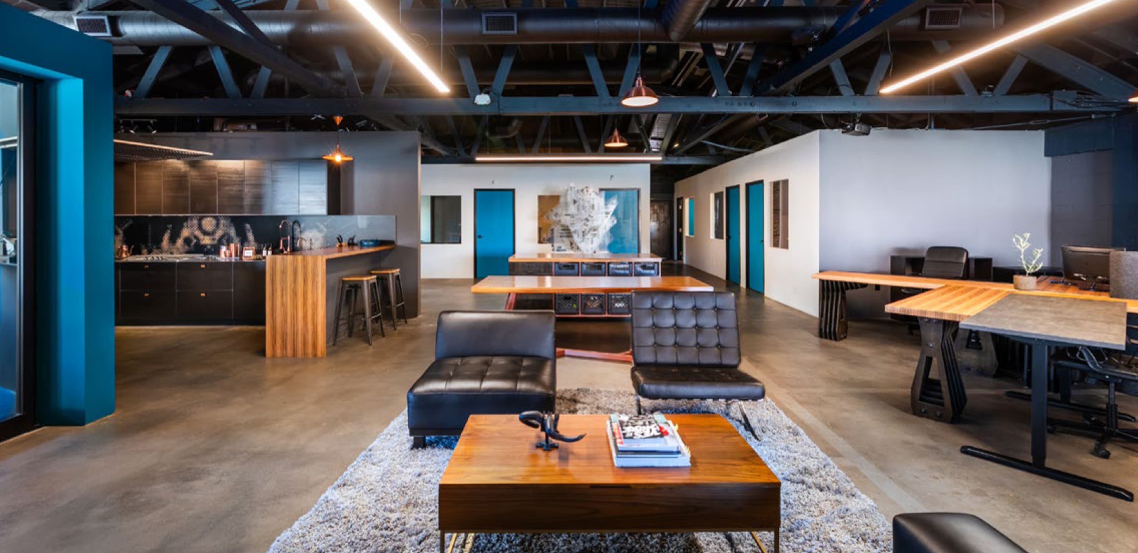 The image shows a modern office space with seating areas, desks, and a kitchenette, featuring industrial design elements like exposed beams and concrete flooring.