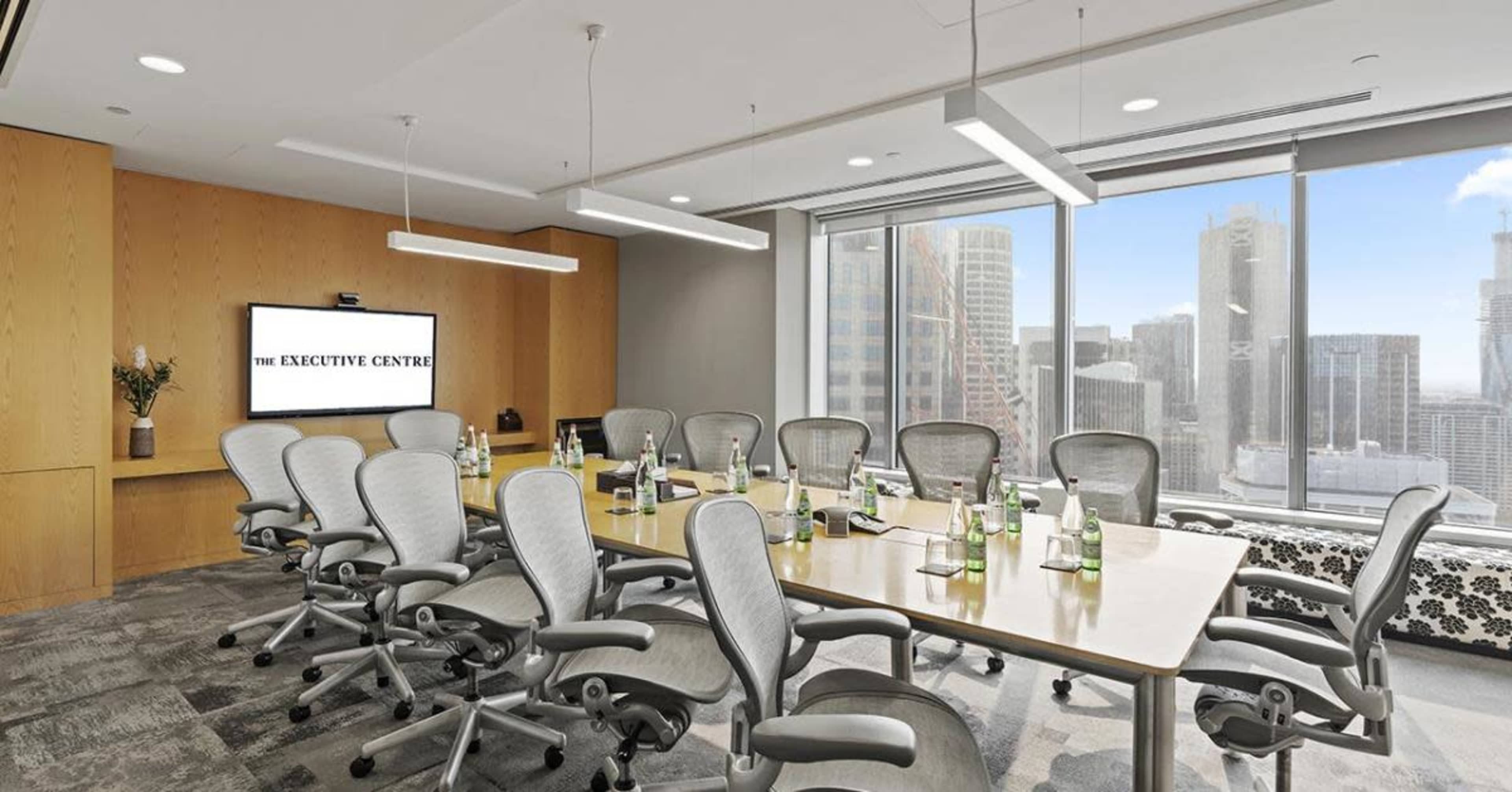 A modern conference room features a long wooden table surrounded by ergonomic chairs, with large windows offering a view of a city skyline.