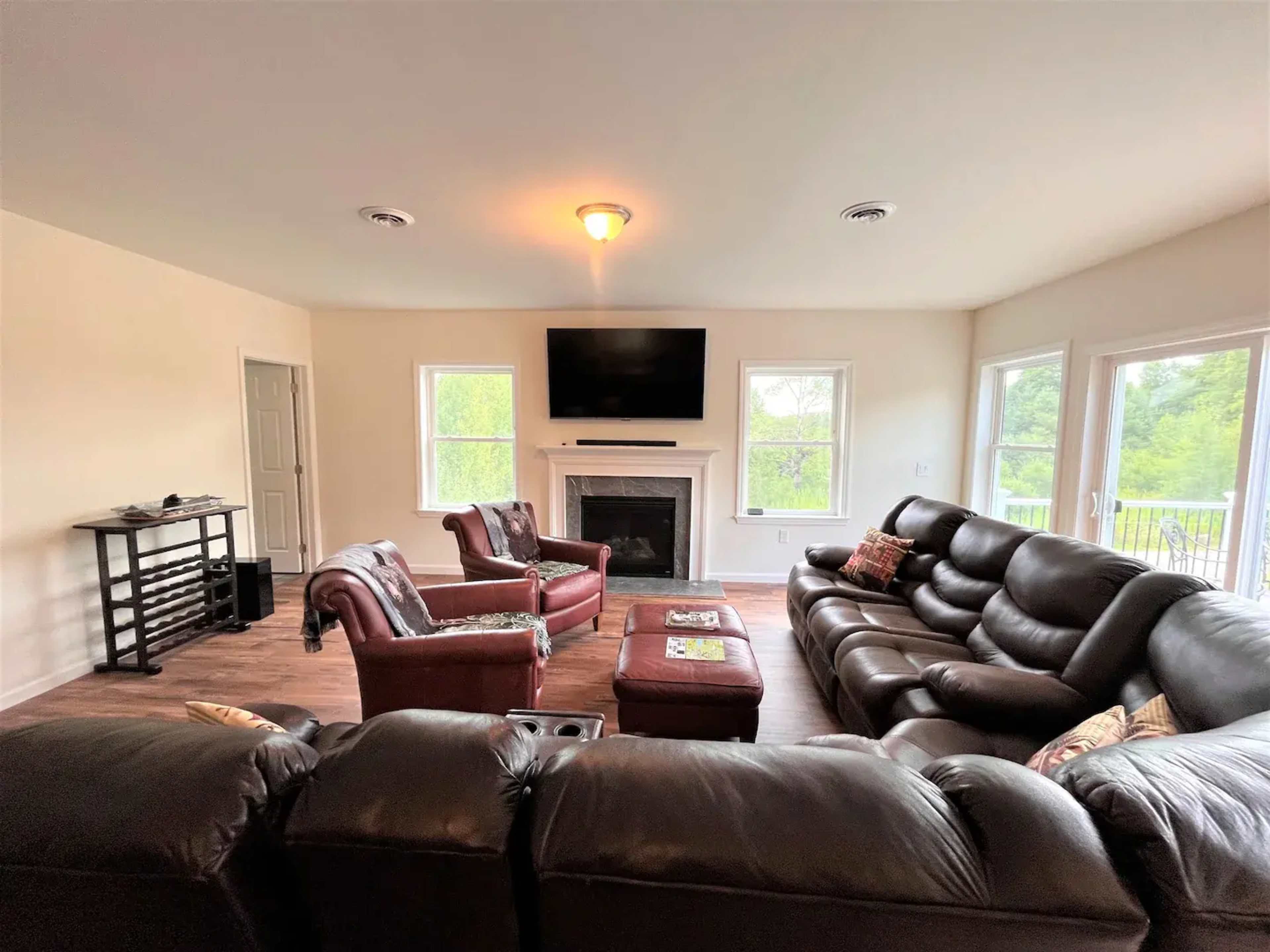 The living room features a dark leather sectional sofa, two matching armchairs, a coffee table, a media console, and a wall-mounted television, with large windows providing views of the outdoors.
