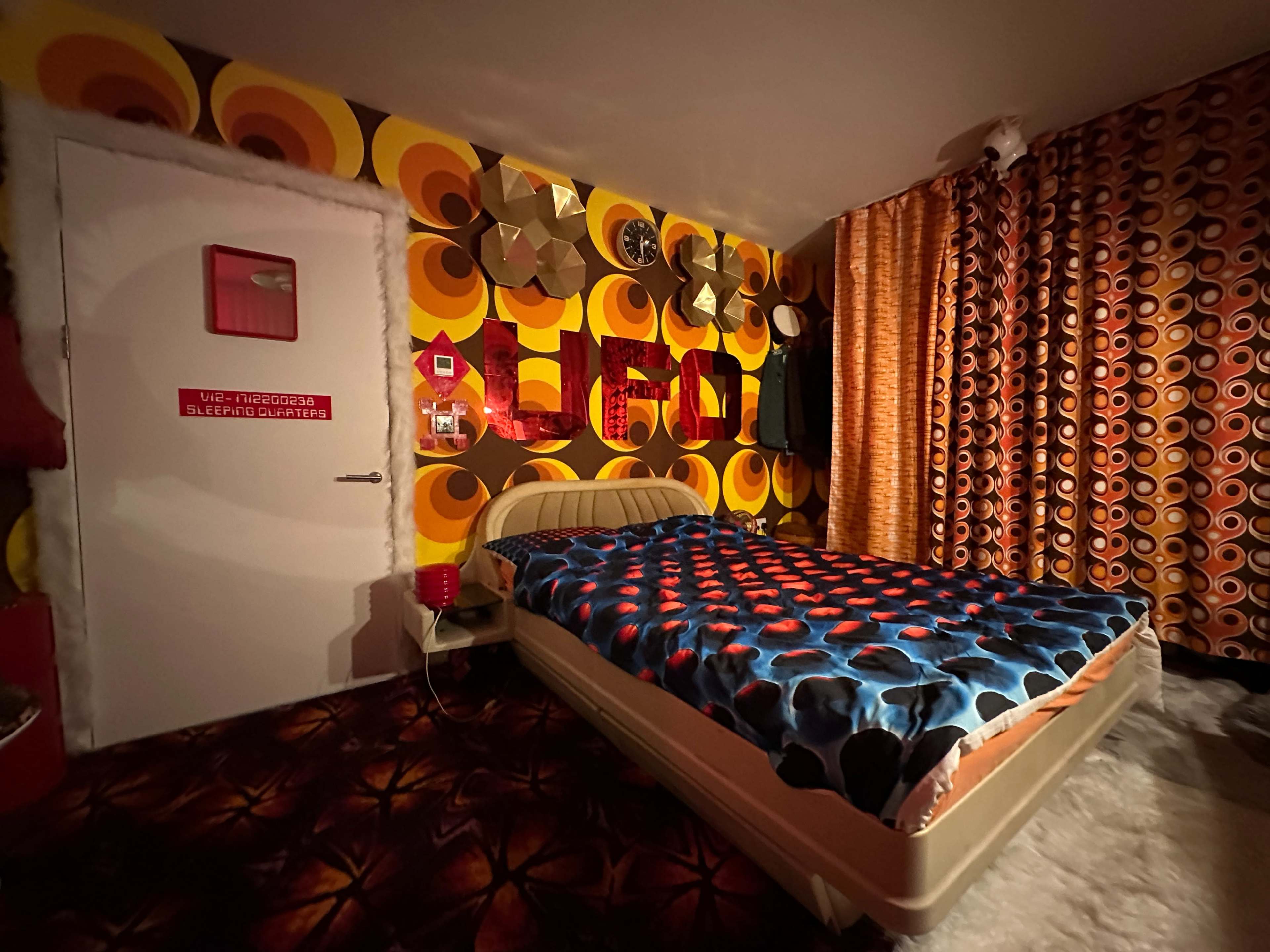 A retro-themed bedroom featuring bold patterned wallpaper, a patterned bedspread, and vibrant curtains.