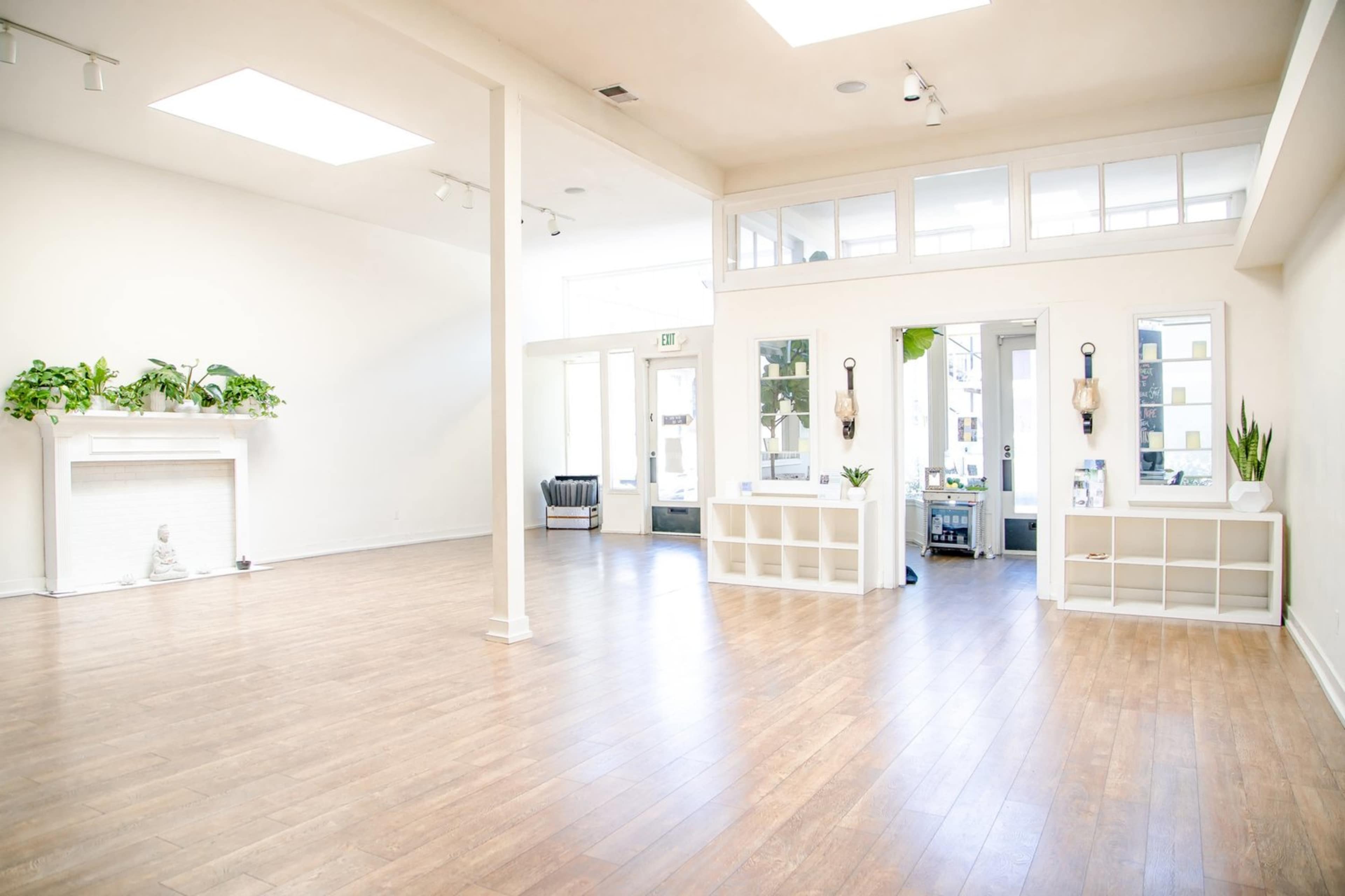 A spacious, empty room with wooden flooring, featuring large windows, minimal furnishings, and plants on display.