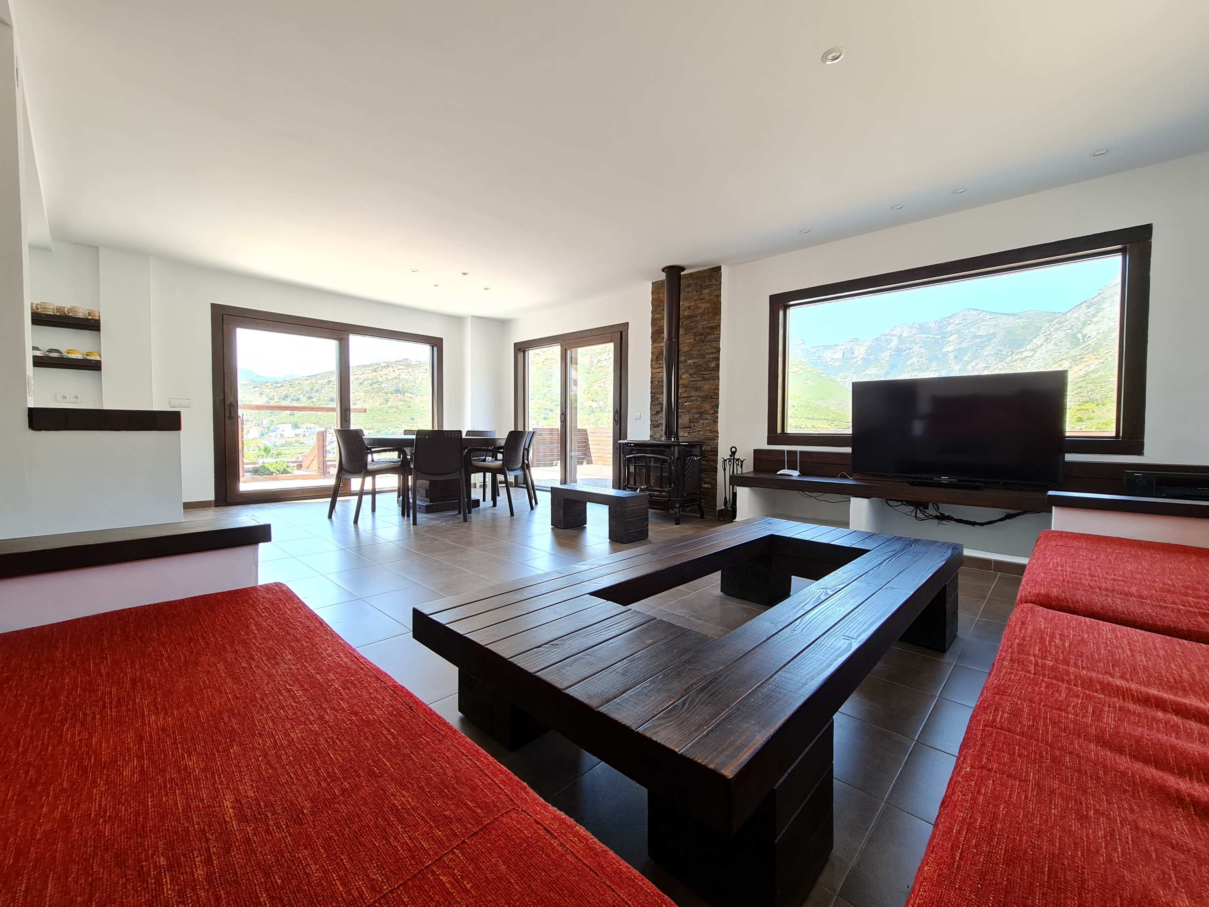 The image shows a modern living room with a wooden coffee table, red seating, a television, and large windows overlooking a mountain view.