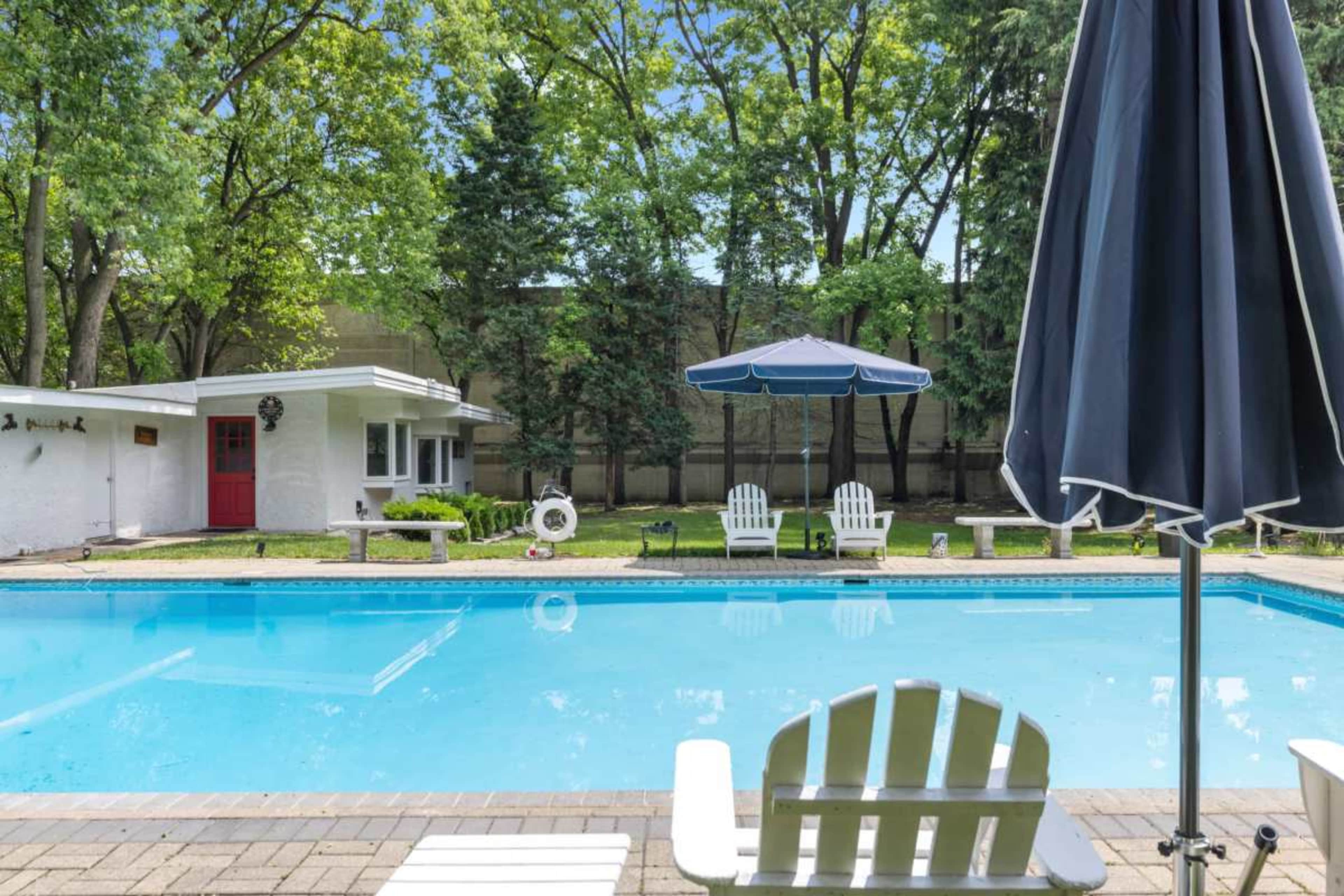 The image shows a swimming pool area with white lounge chairs, a blue umbrella, and a small building in the background surrounded by trees.
