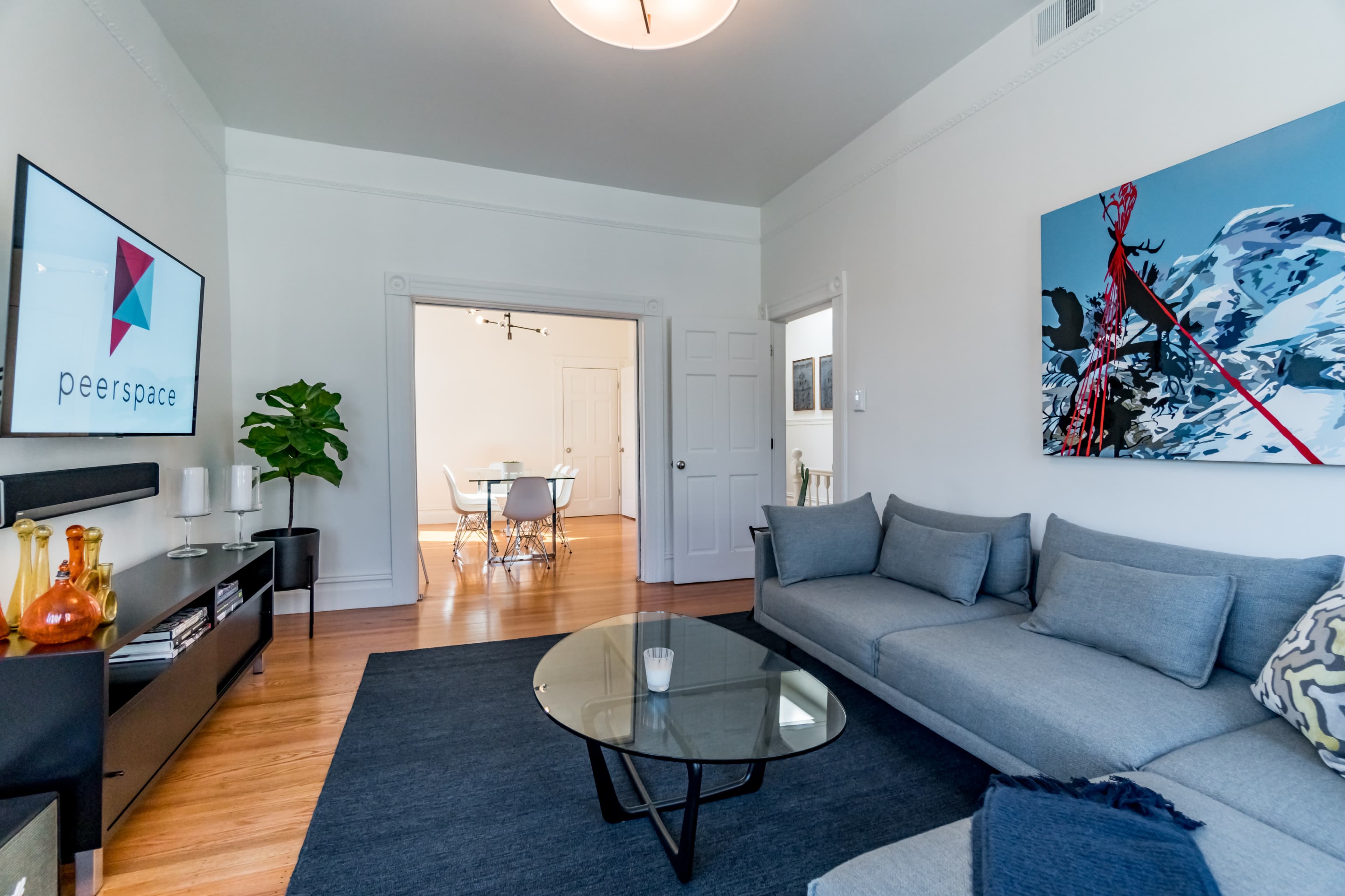 A modern living room with a gray sectional sofa, a glass coffee table, and a television displaying the Peerspace logo, leading to an open dining area in the background.