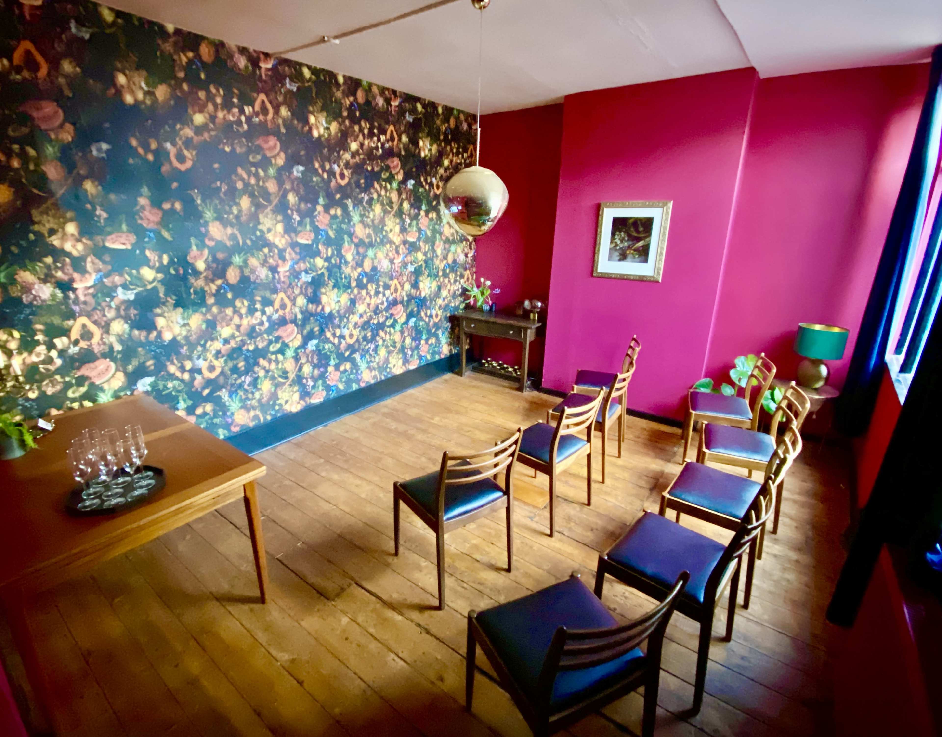 A small room features a floral patterned wall, a wooden table with a glassware set, and several wooden chairs arranged in a semi-circle.