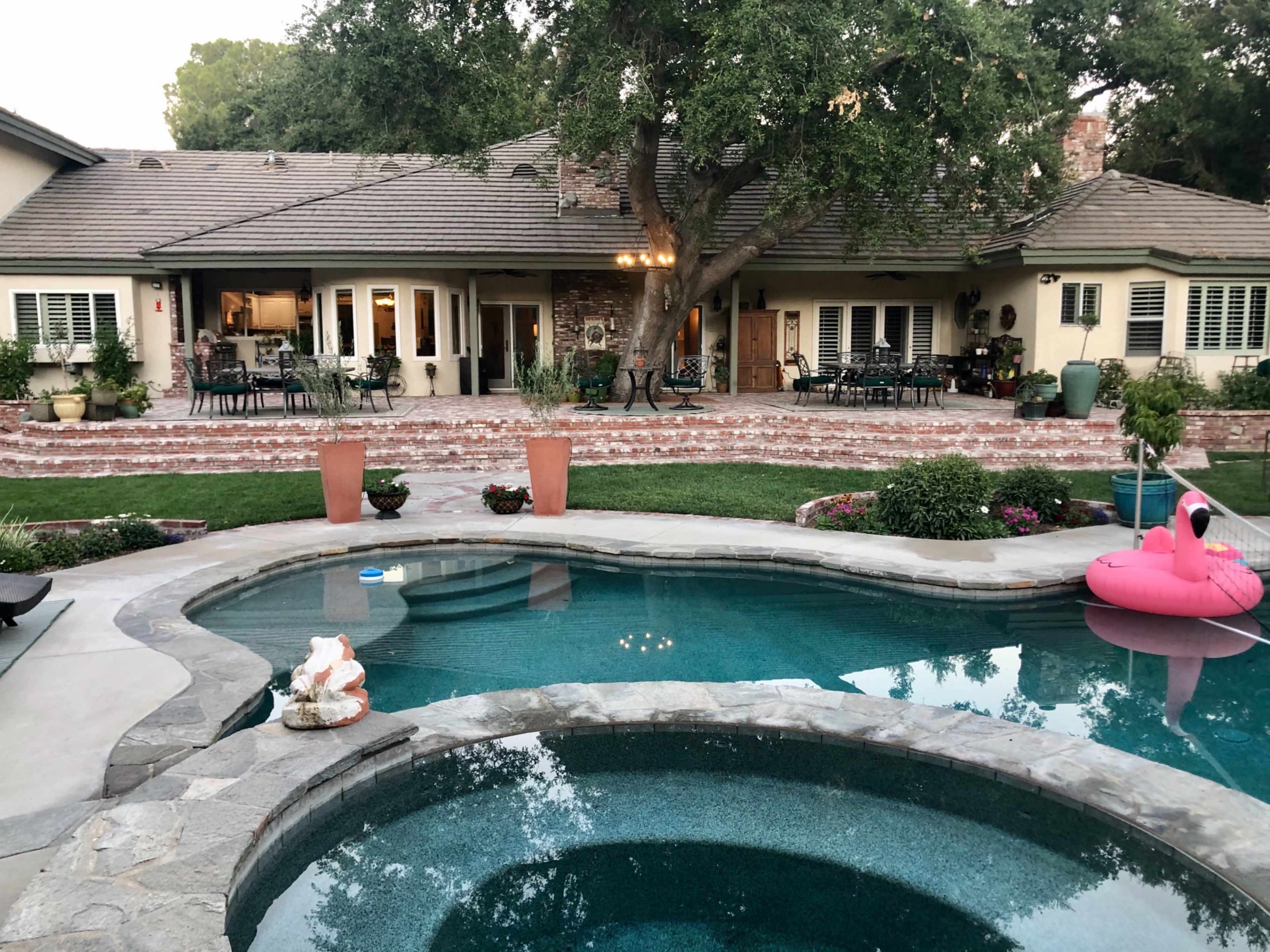 A landscaped backyard with a swimming pool, a spa, and a large house in the background surrounded by greenery and patio furniture.