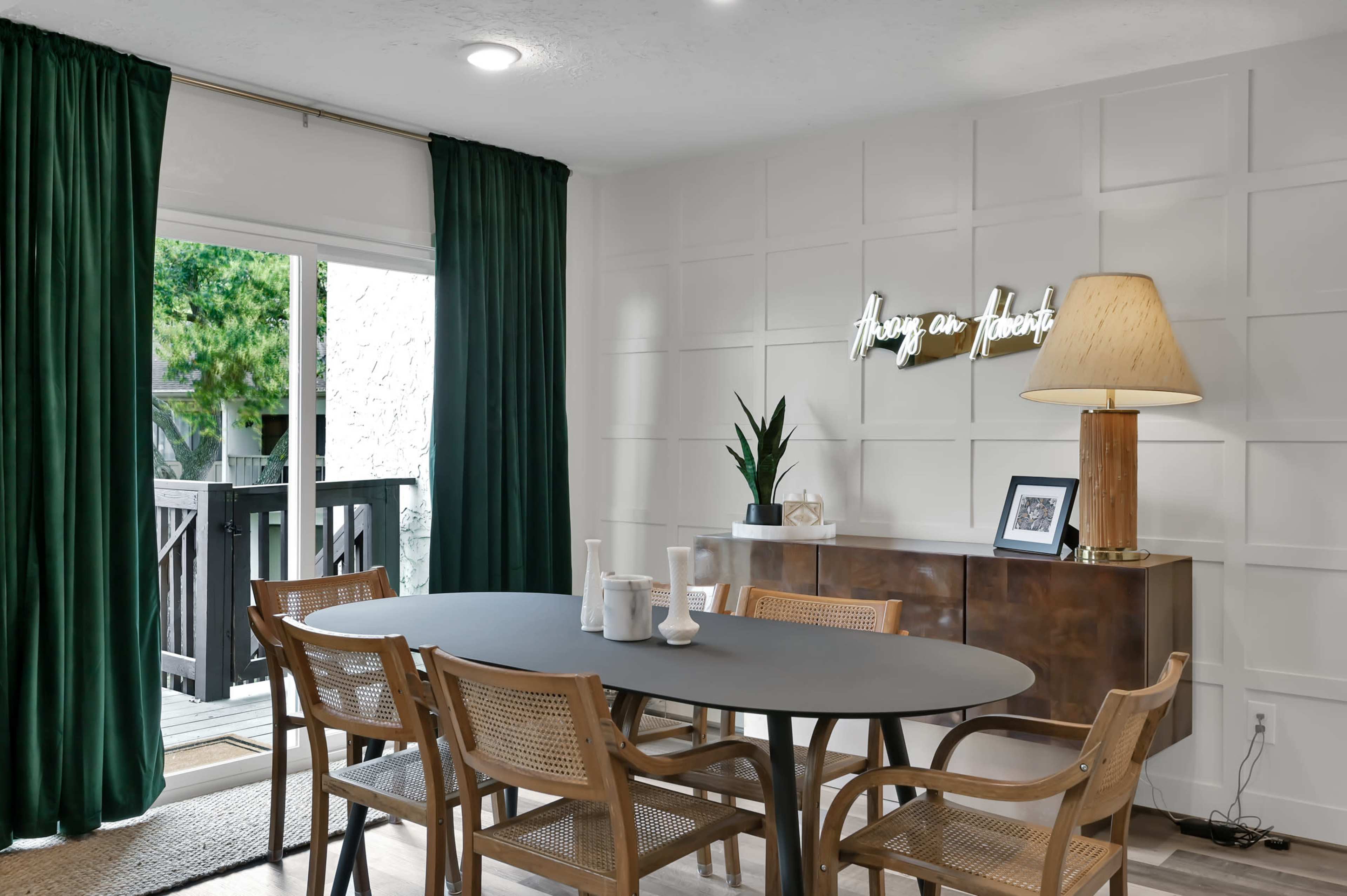 A dining area features a round table surrounded by wooden chairs, with a green curtain, a decorative lamp, and a console table against a paneled wall.